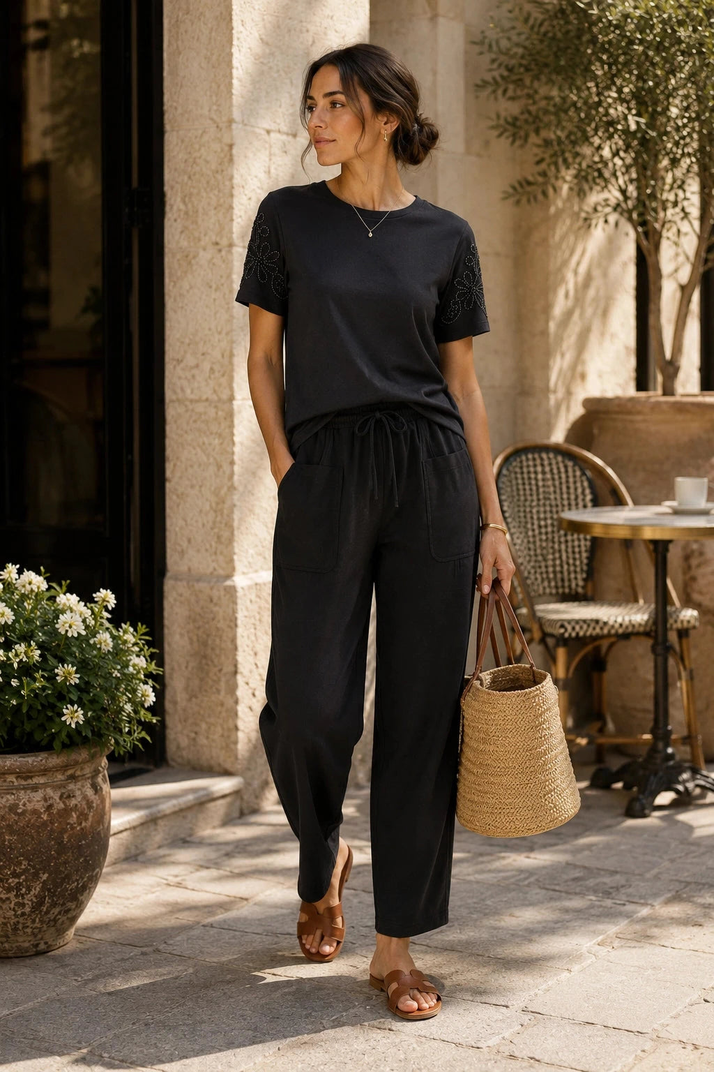 A woman stands on a sunlit stone walkway wearing a black short-sleeve top with textured sleeves and matching black drawstring pants. She is holding a woven straw tote bag and wearing brown sandals, with a cafe table and potted plants in the background.