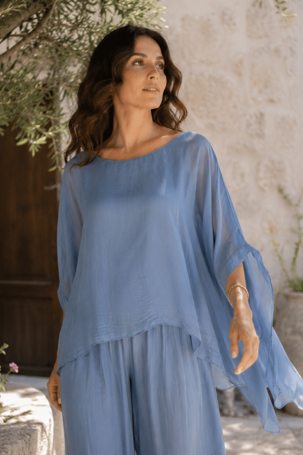 A woman wearing a light blue two-piece set consisting of a loose, semi-sheer long-sleeve top and matching wide-leg pants, standing outdoors against a rustic stone wall.