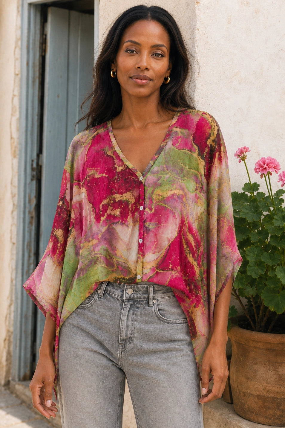 A woman wearing a loose, semi-sheer button-up blouse with a vibrant pink, green, and yellow floral abstract print, paired with light-wash gray jeans, standing outdoors in front of a rustic white wall and potted flowers.