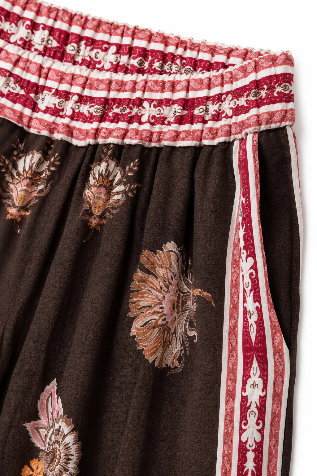 Close-up of a skirt with a dark brown base, featuring large beige and pink floral prints, a red and white patterned waistband, and a red side stripe with white motifs.