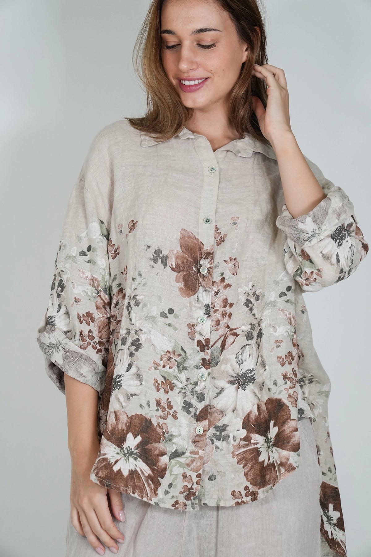 A woman wearing a loose, long-sleeve button-up blouse with a brown and beige floral print, paired with a matching light-colored skirt, set against a plain light gray background.