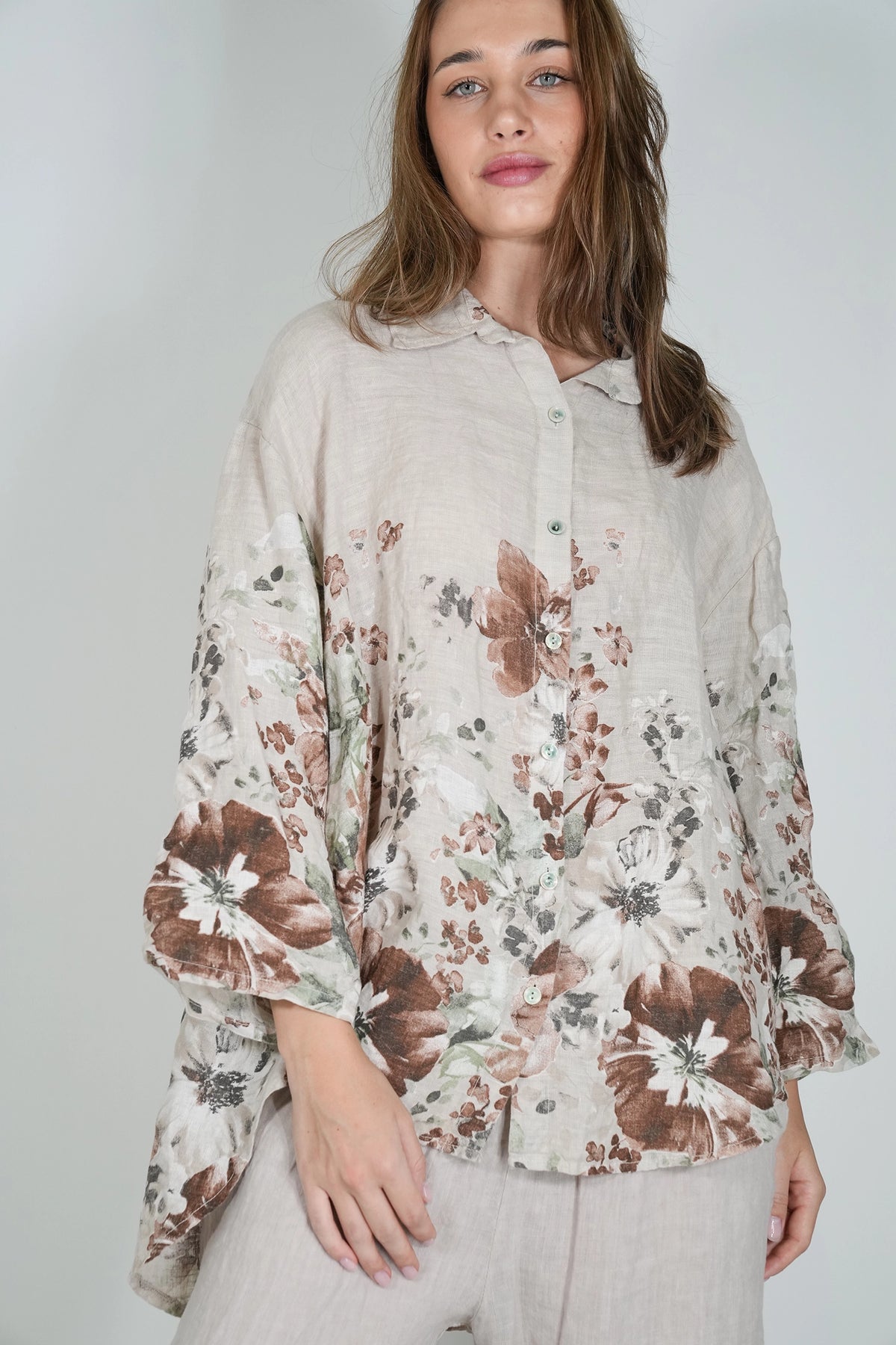 A woman wearing a light beige linen button-up blouse with a brown and white floral print, paired with light beige trousers, against a plain light gray background.