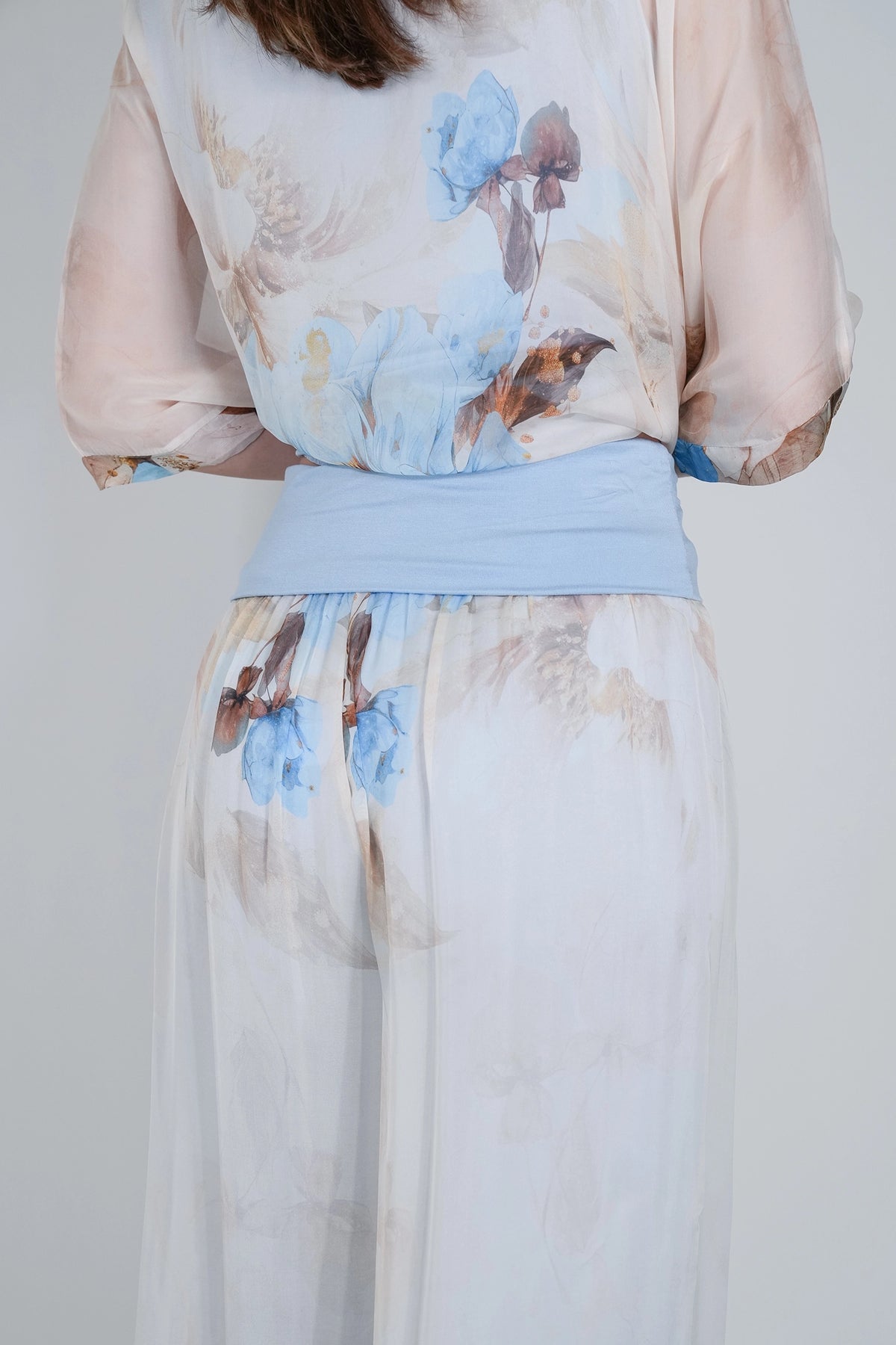 A close-up of a model from the back wearing a sheer, light peach and white floral jumpsuit with a light blue waistband.