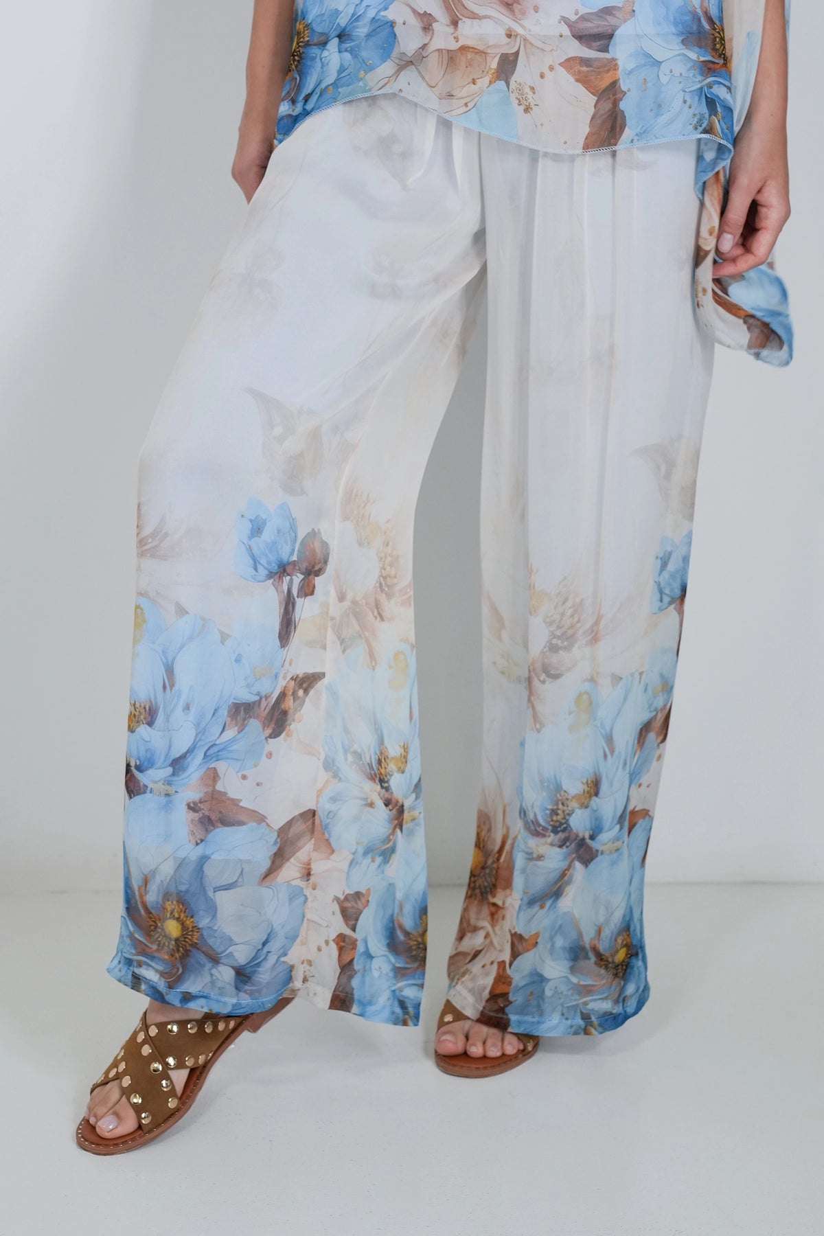 A person wearing wide-leg white pants with a blue and beige floral print, paired with brown studded sandals, shown from the waist down against a plain light background.