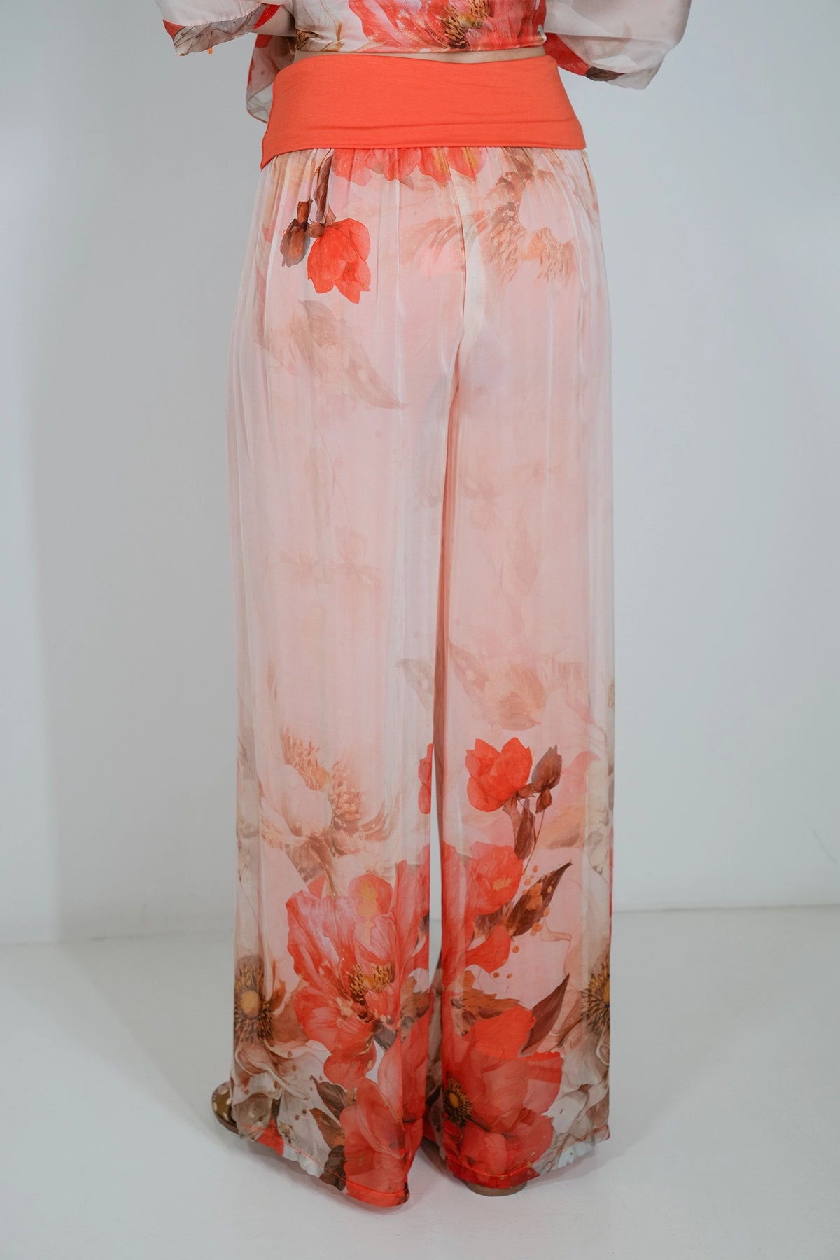A person wearing sheer peach and orange wide-leg pants with a large floral print in red, orange, and brown, paired with a solid orange waistband and matching top.
