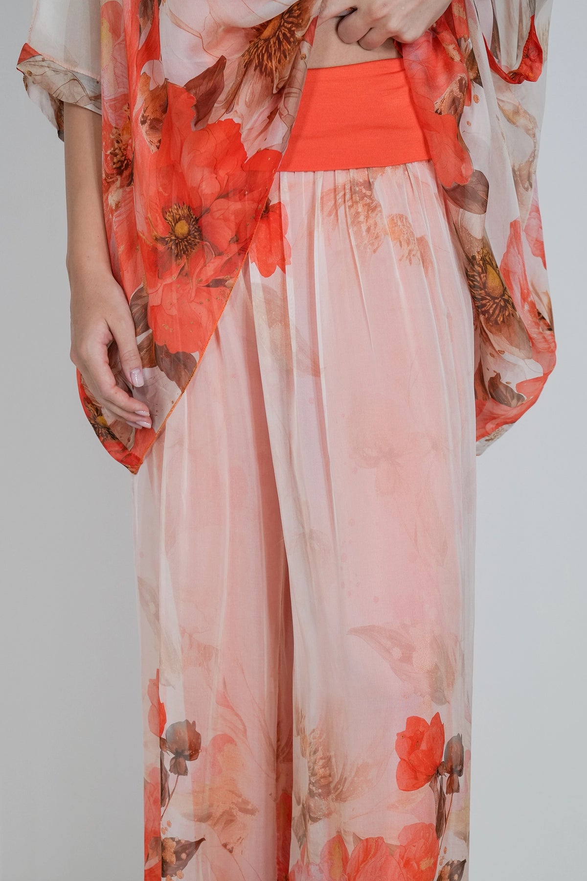 A close-up of a person wearing sheer peach and coral floral pants with a matching orange waistband and a sheer floral kimono-style top draped over one shoulder, against a light gray studio background.