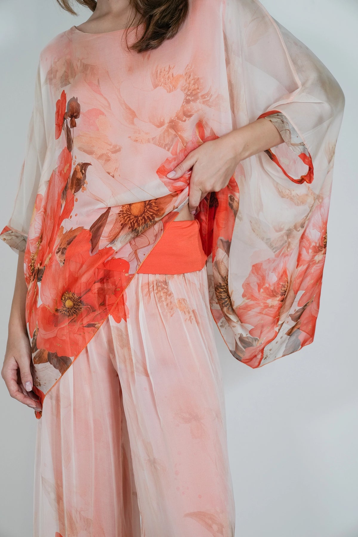 A woman wearing a sheer peach and coral floral jumpsuit or matching set with wide flowing sleeves and a matching sheer wrap, photographed against a plain light background.