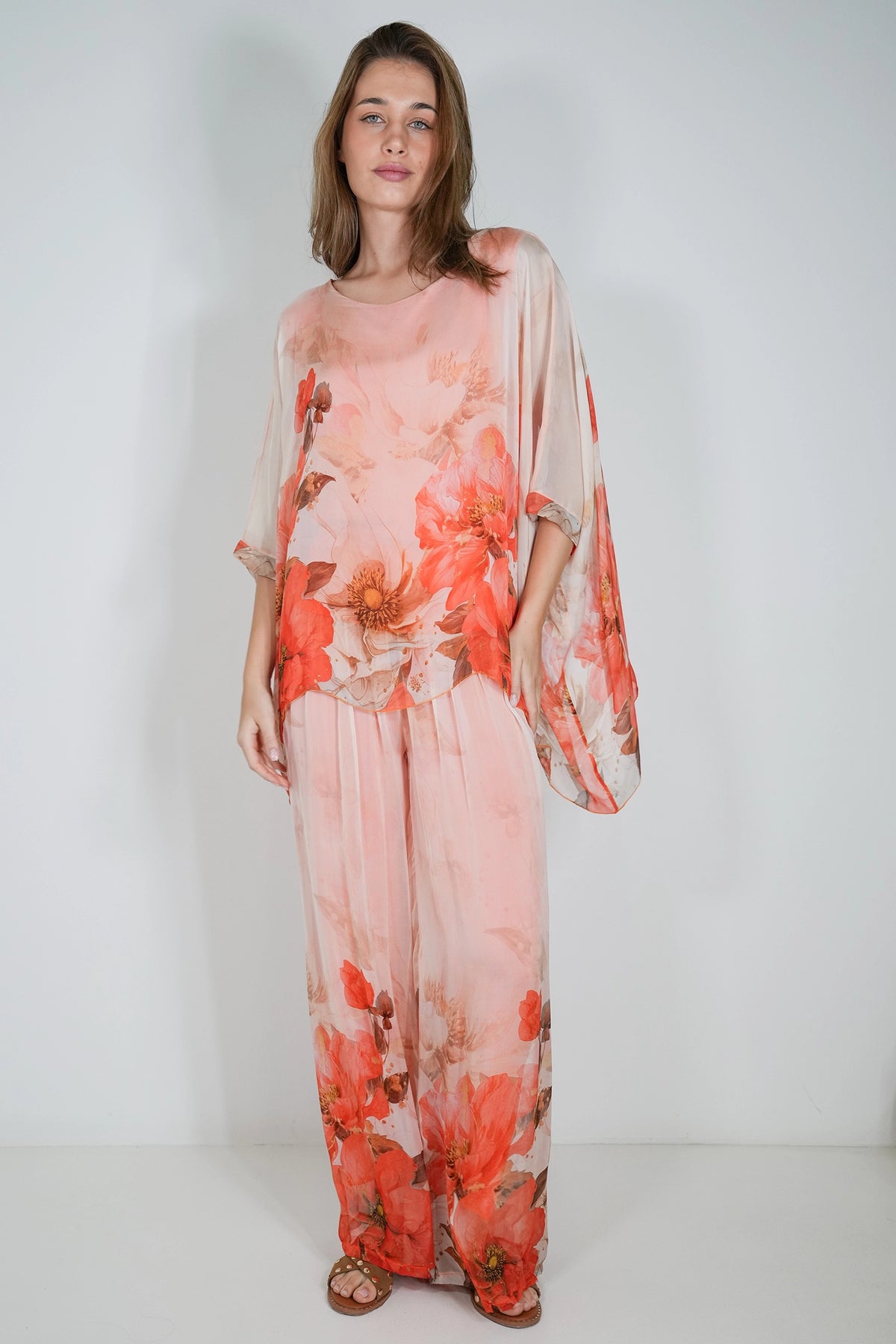 A woman modeling a light pink two-piece set with a large red and orange floral print, featuring a loose kaftan-style top with wide sleeves and matching wide-leg pants, set against a plain white studio background.