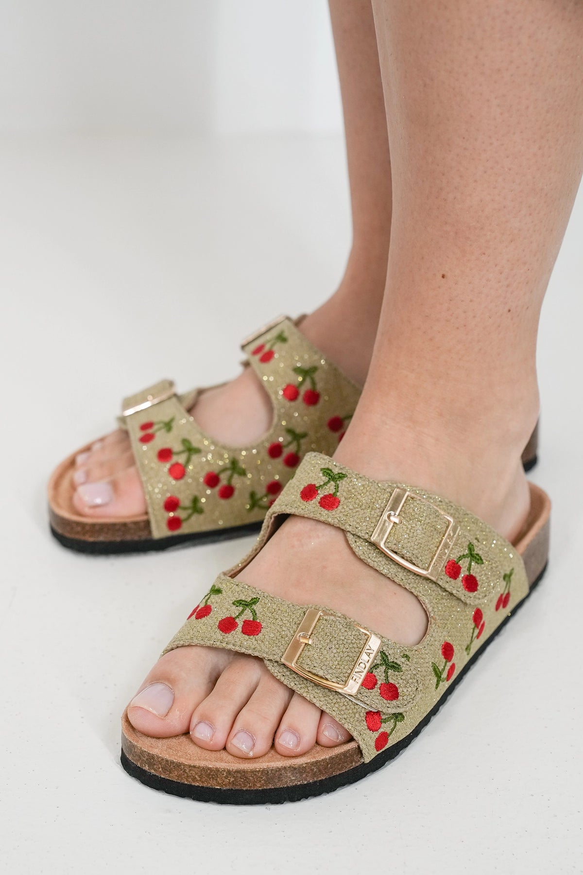 A pair of double-strap slide sandals worn on feet, featuring a beige glittery fabric upper with an embroidered cherry pattern and gold-tone buckle straps, set against a clean white background.