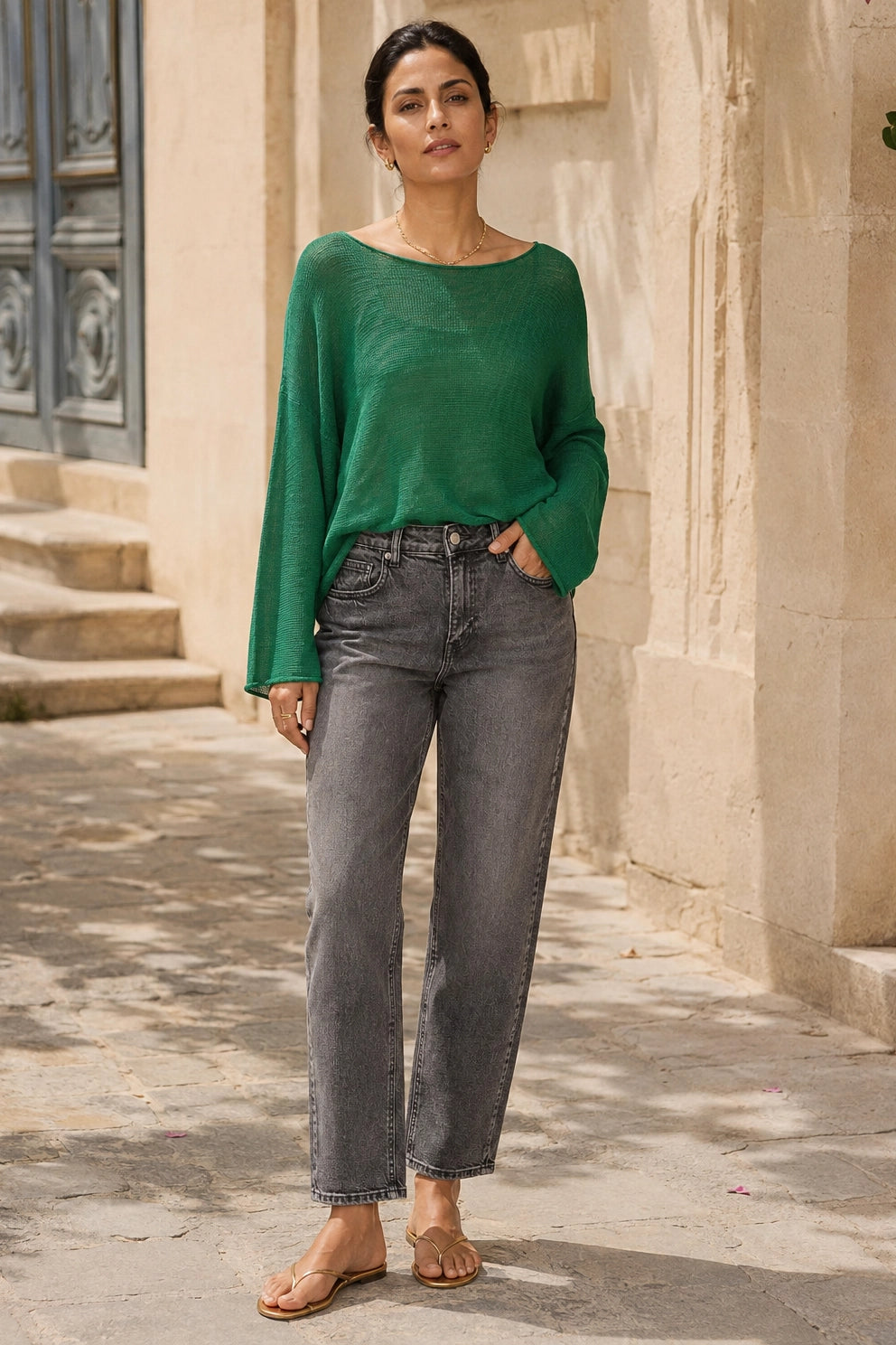 A woman stands outdoors wearing a loose, long-sleeve green knit top and dark gray straight-leg jeans, with tan sandals, posed in front of a beige stone building.