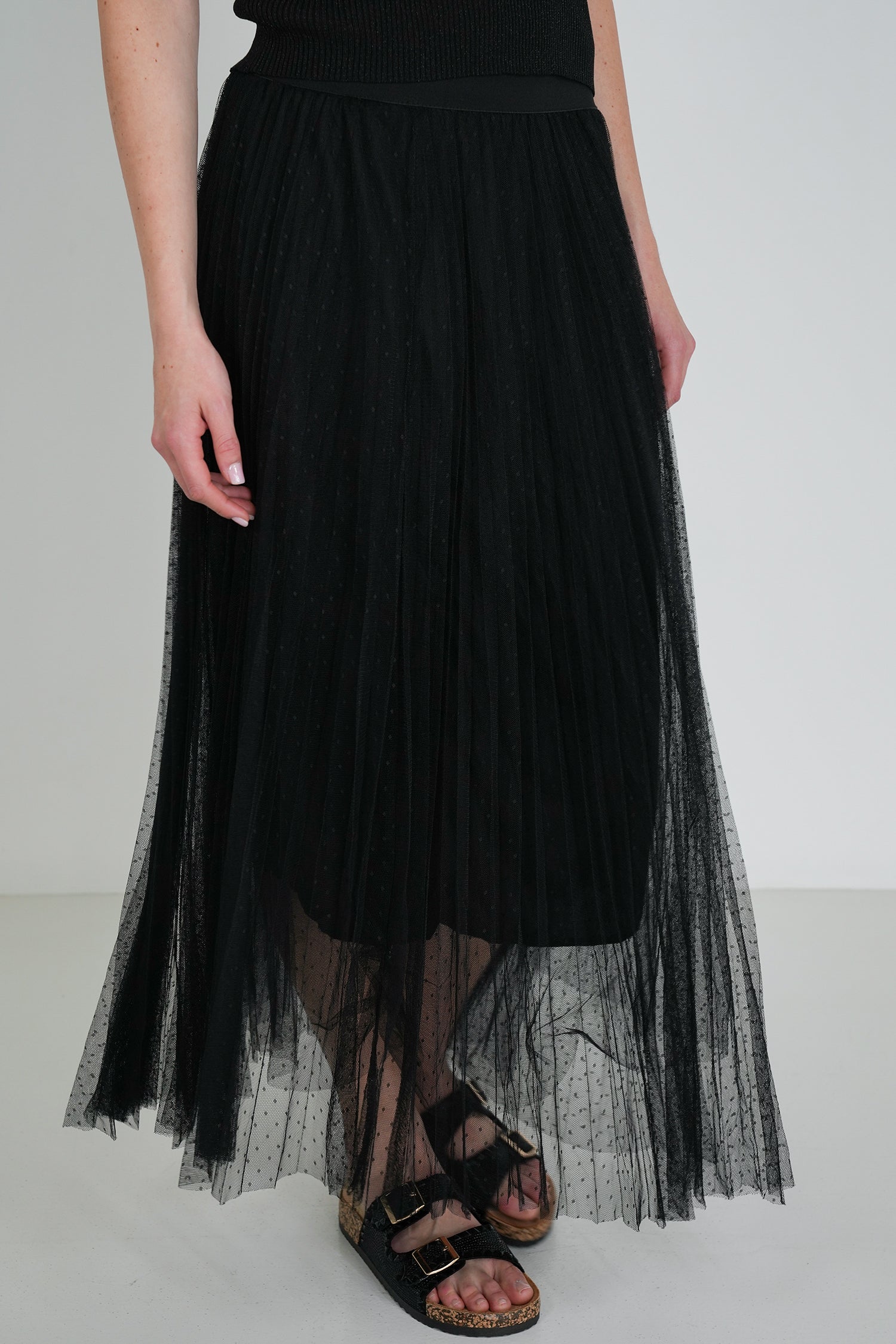 Close-up of black pleated tulle midi skirt with sheer overlay, showing waist detail and black sandals with gold buckles.