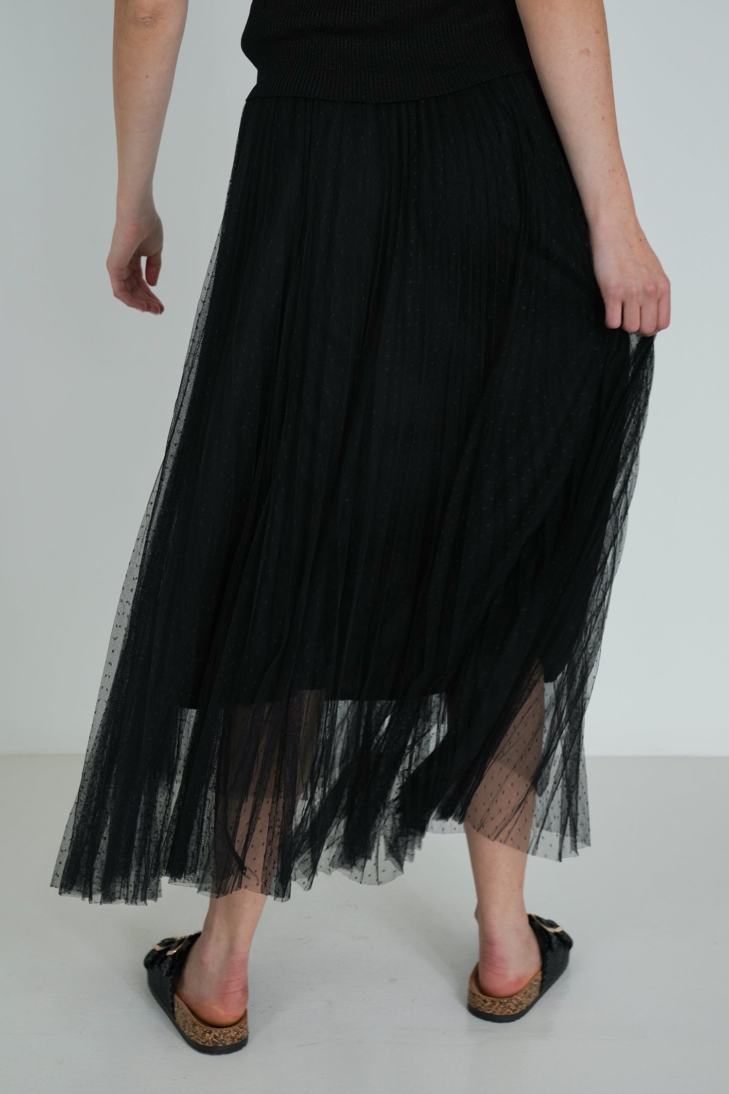Back view of black pleated tulle midi skirt with sheer overlay, showing flowy movement and styled with black sandals.