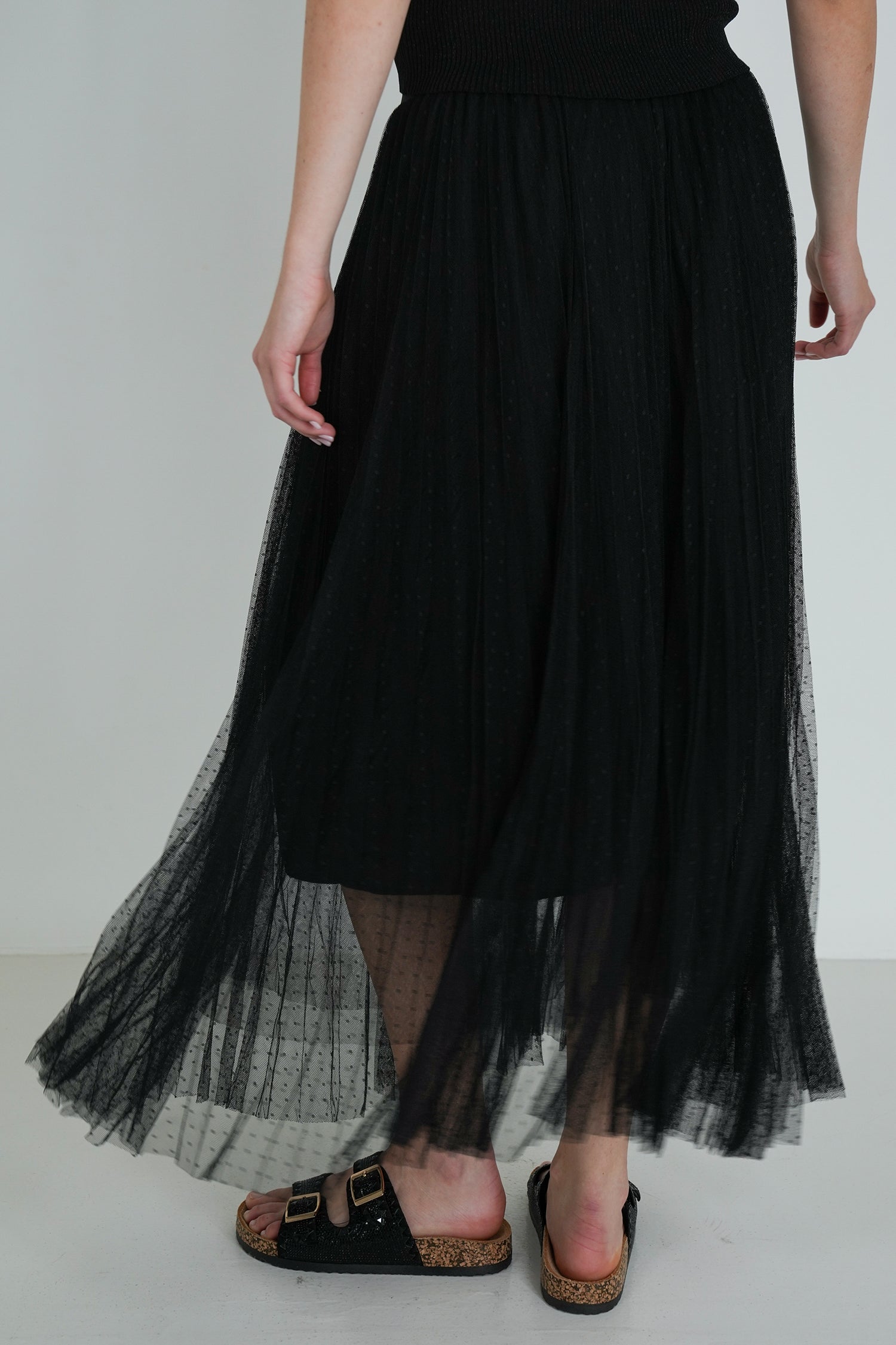 Back view of black pleated tulle midi skirt with sheer layers, showing flowy fabric detail and black sandals with gold buckles.