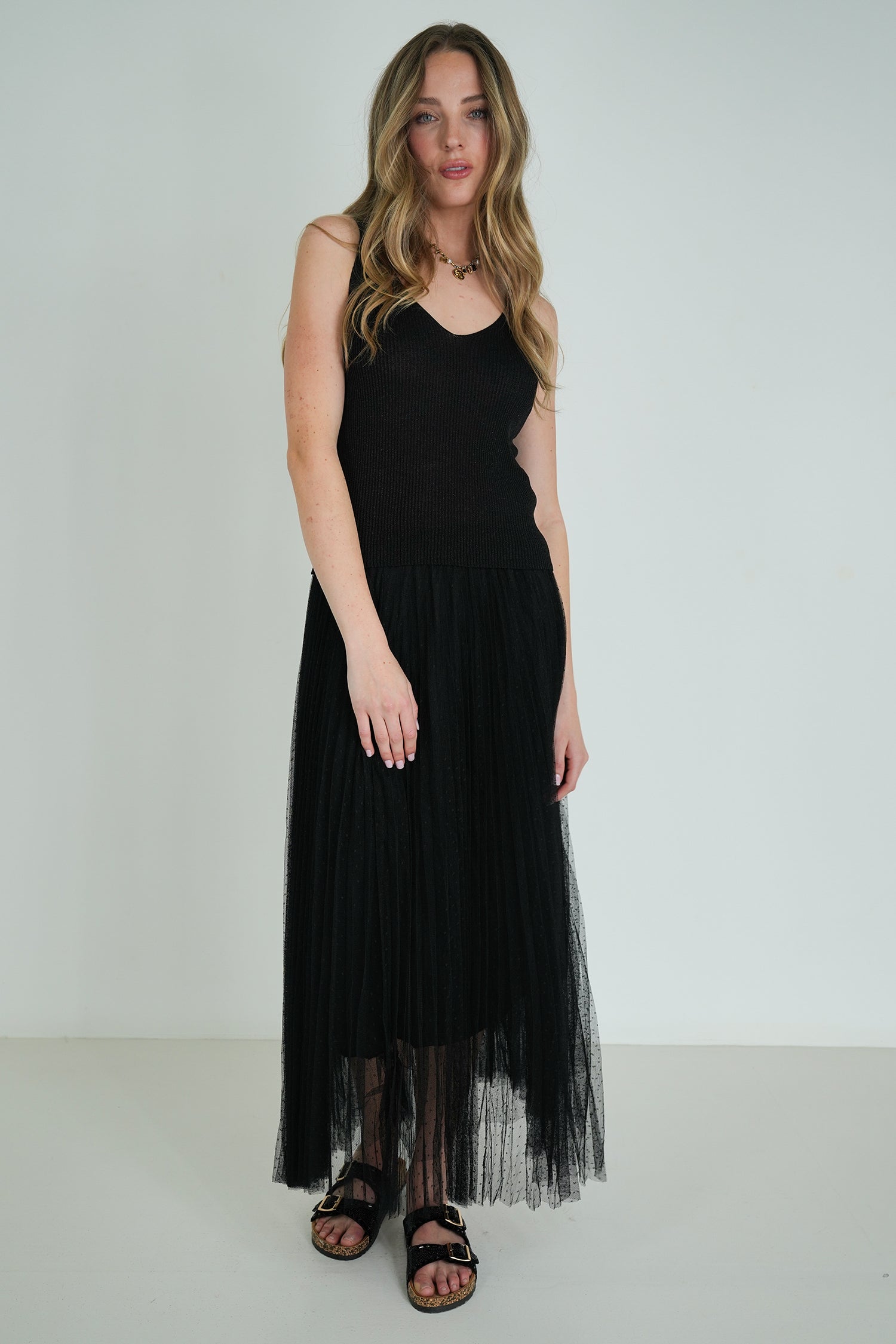 Model wearing a sleeveless black ribbed top and pleated tulle midi skirt, paired with black sandals, posing against a white wall.