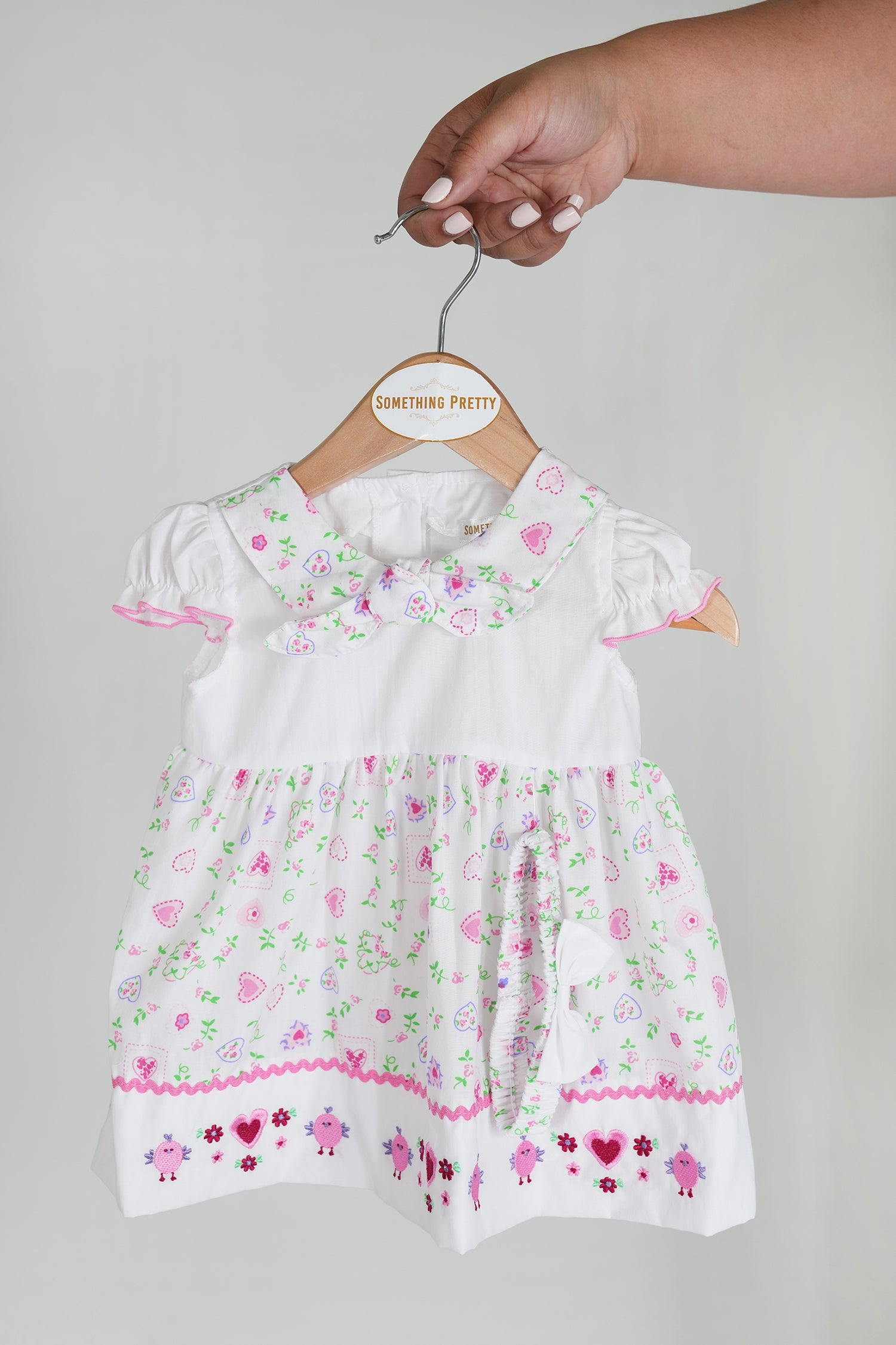 A white peggy dress with short sleeves and decorative elements in pink and green, hanging on a wooden hanger.