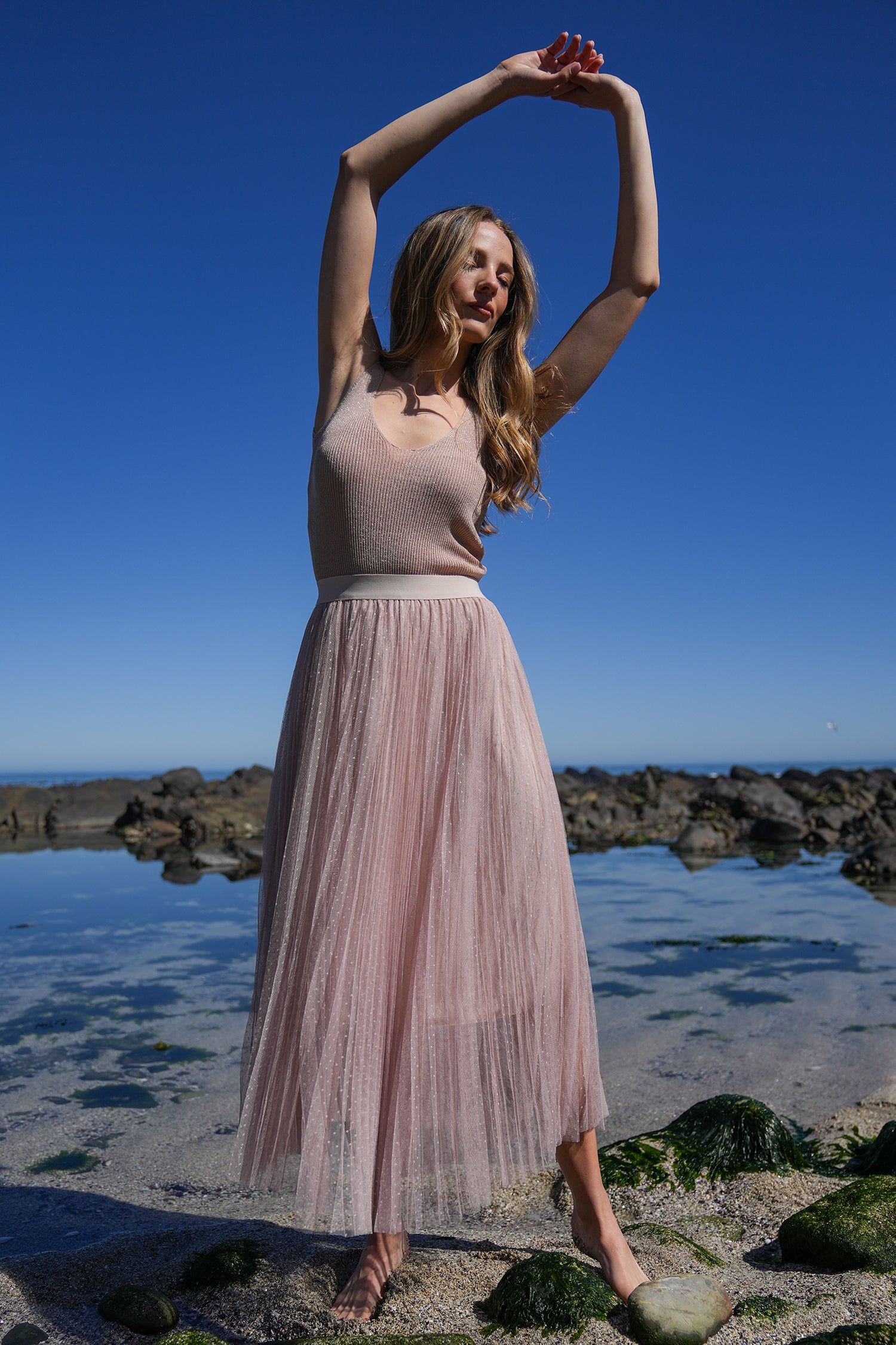 Model wearing a blush pink pleated tulle midi skirt and beige top, posing outdoors near rocks under clear blue sky.