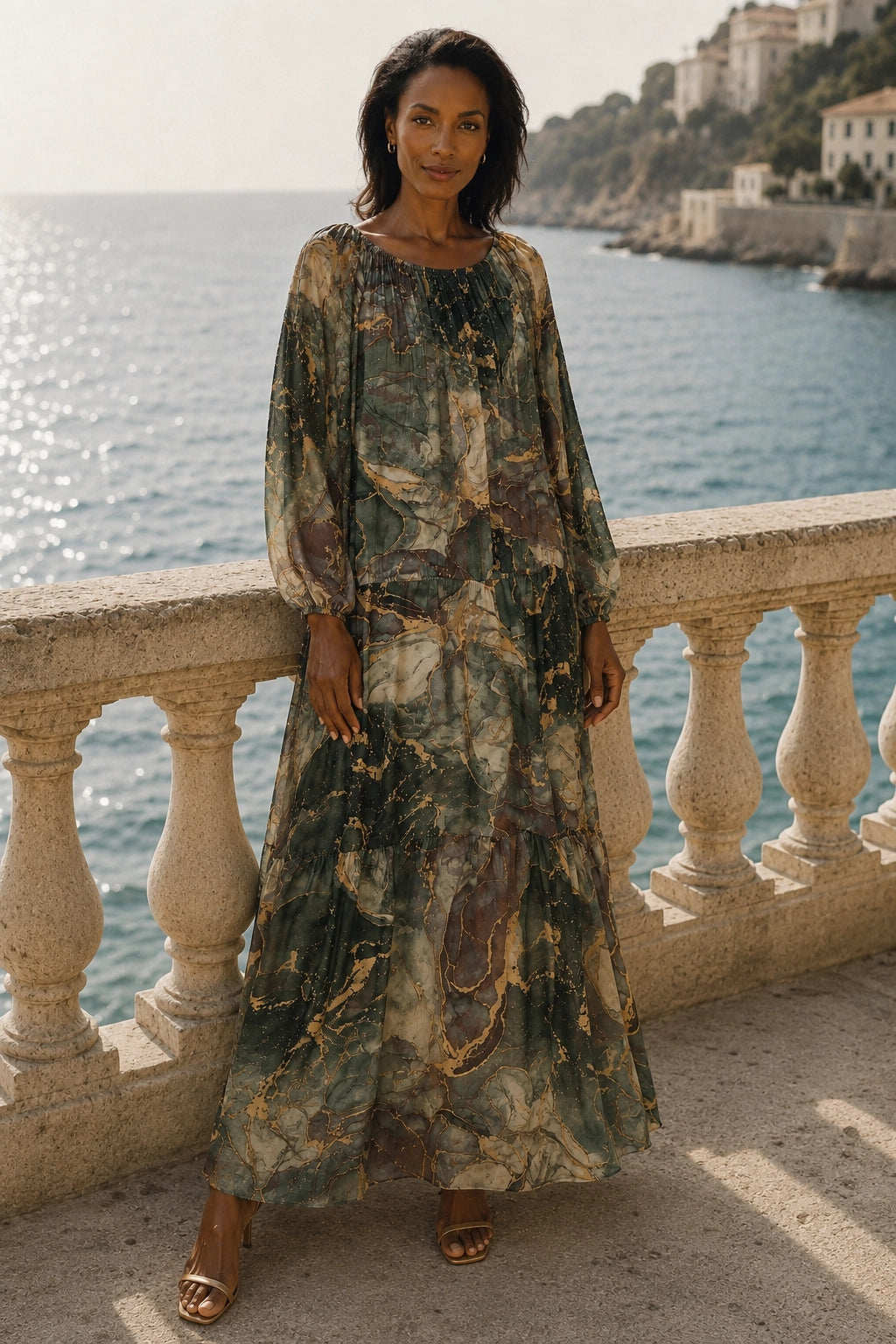 A woman stands on a stone balcony overlooking the sea, wearing a long-sleeved maxi dress with an abstract marbled pattern in olive green, beige, and brown tones.