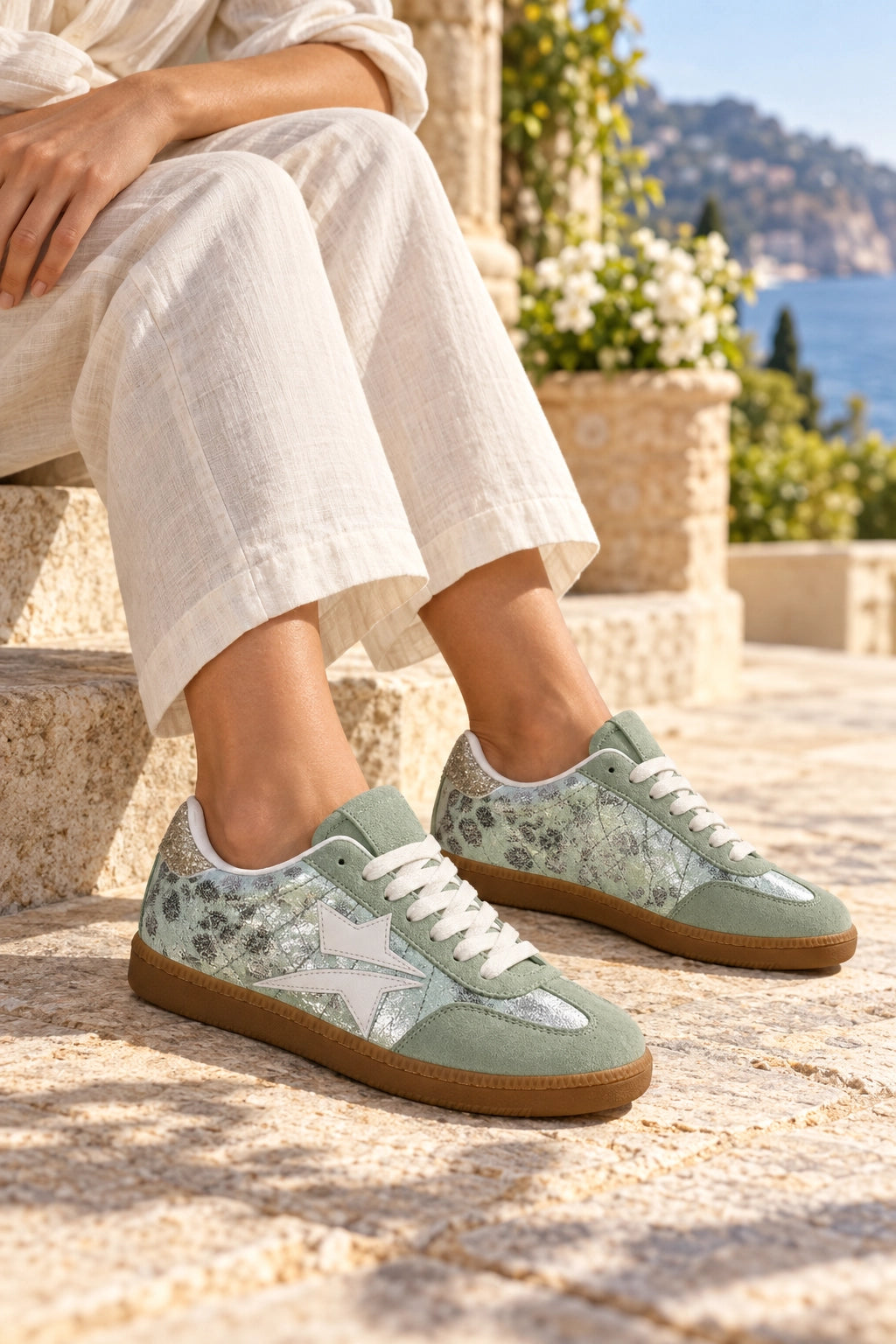 A person sitting outdoors on stone steps wearing light sage green low-top sneakers with white star details, white laces, and a floral pattern on the sides, paired with cream linen pants. A sunny coastal landscape with blue water and greenery is visible in the background.
