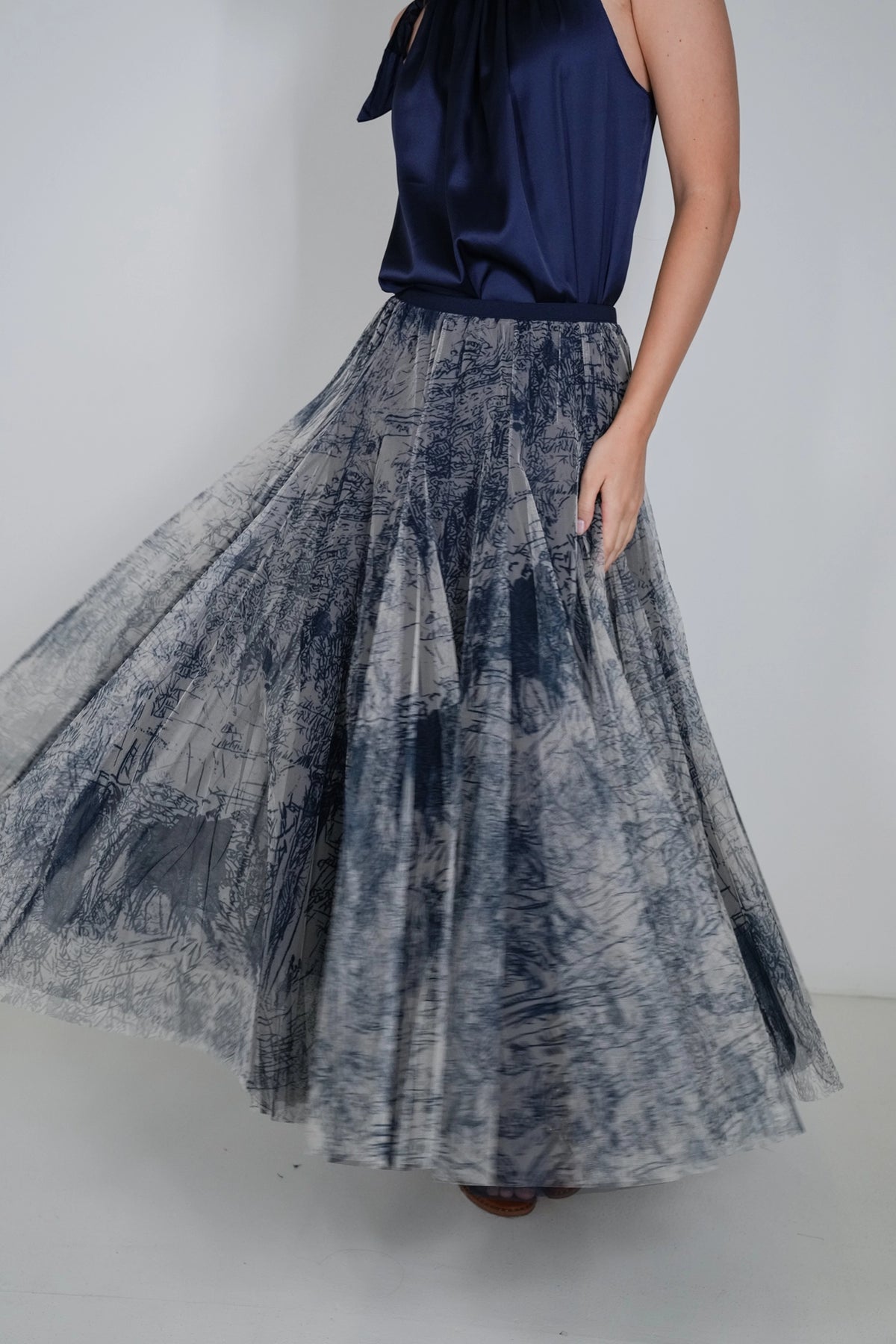 A woman wearing a navy blue satin halter top and a full, flowing pleated skirt with an abstract blue, gray, and white brushstroke pattern, captured against a plain light gray studio background.