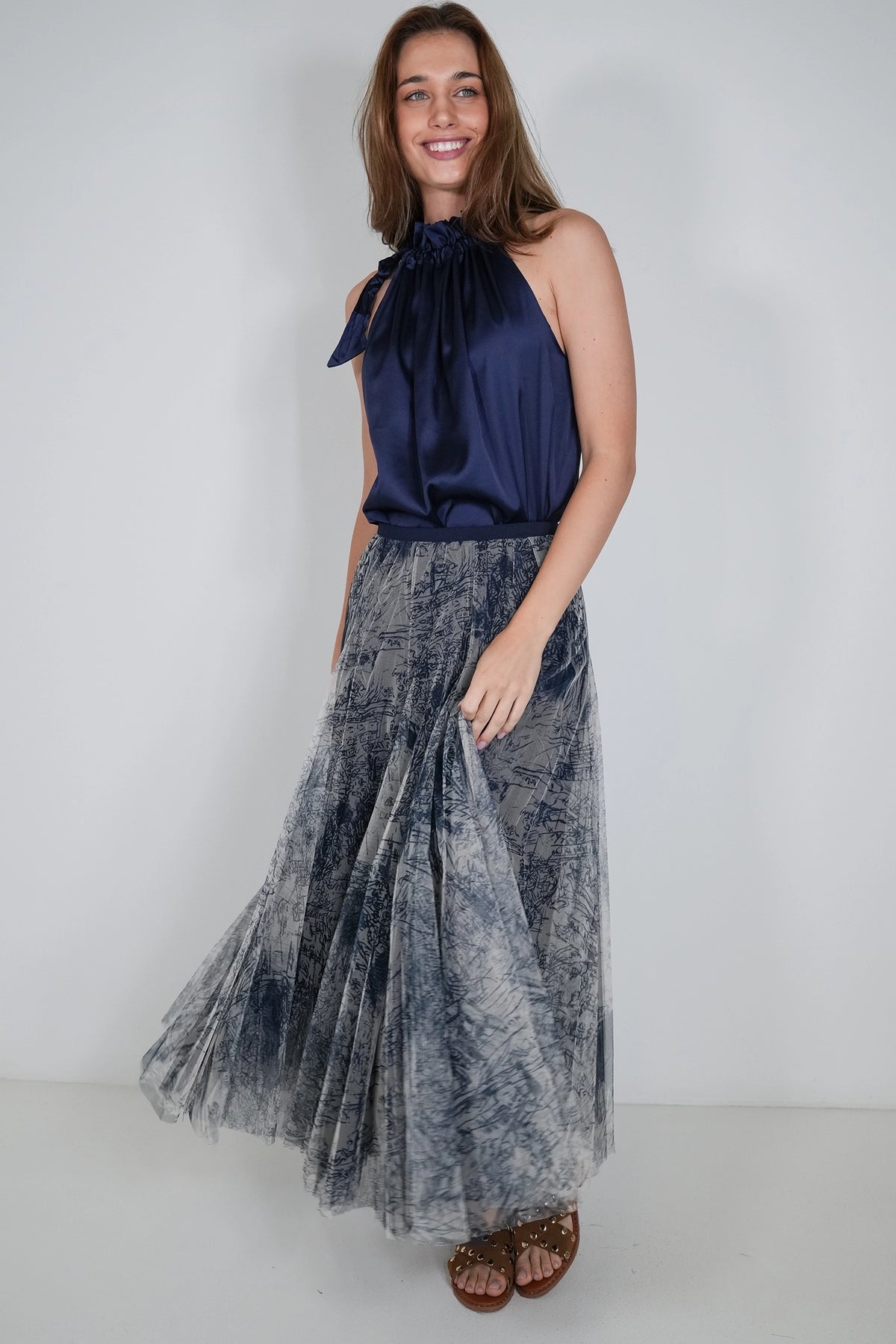 A woman modeling a sleeveless halter-neck top in dark navy blue satin paired with a long pleated tulle skirt in a gray and blue tie-dye pattern, standing against a plain light gray studio background.