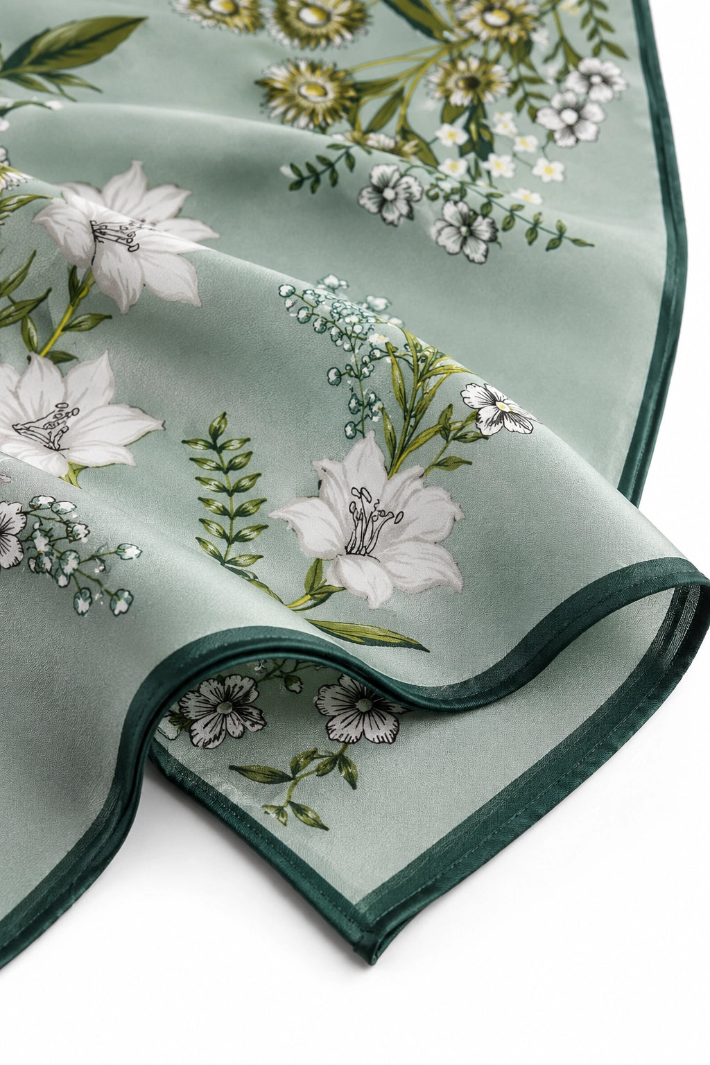 A close-up of a folded dusty sage green scarf featuring a delicate botanical print of white lilies, green leaves, and small yellow flowers, finished with a dark green border.