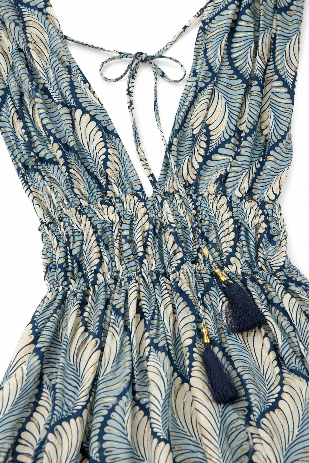 Close-up of a sleeveless dress with a deep V-neckline, tie-back detail, gathered waist, and navy blue tassel accents on a cream and navy patterned fabric.