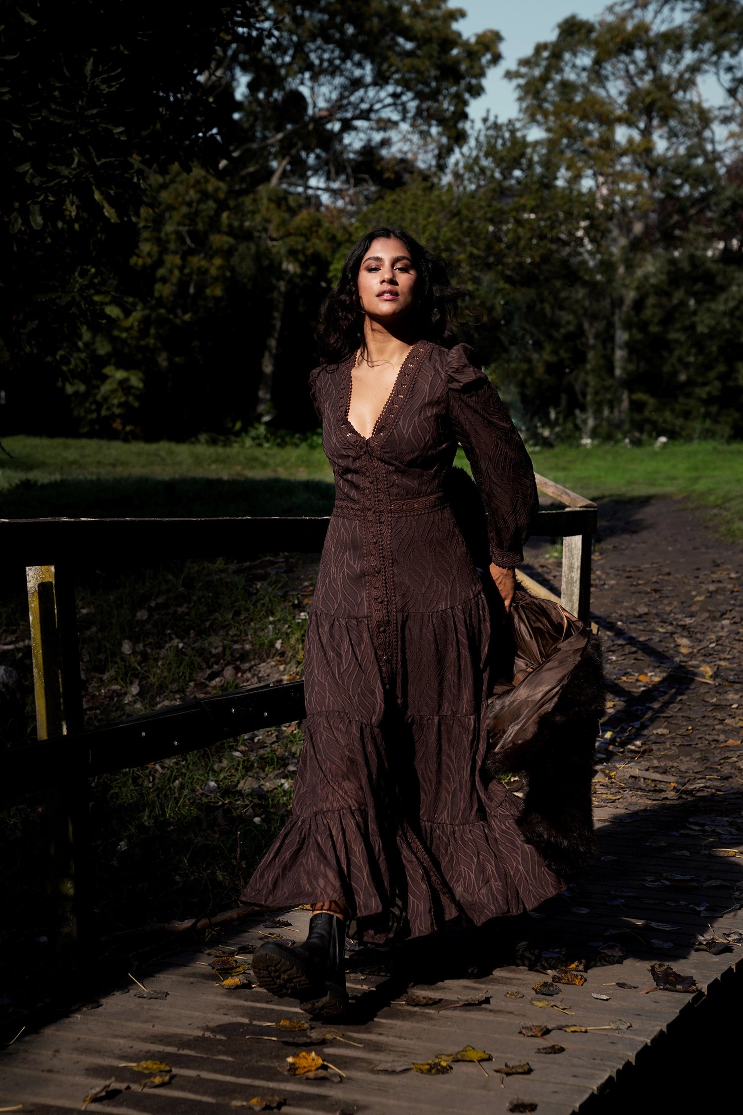 Woman in a brown dress standing on a wooden bridge with trees in the background