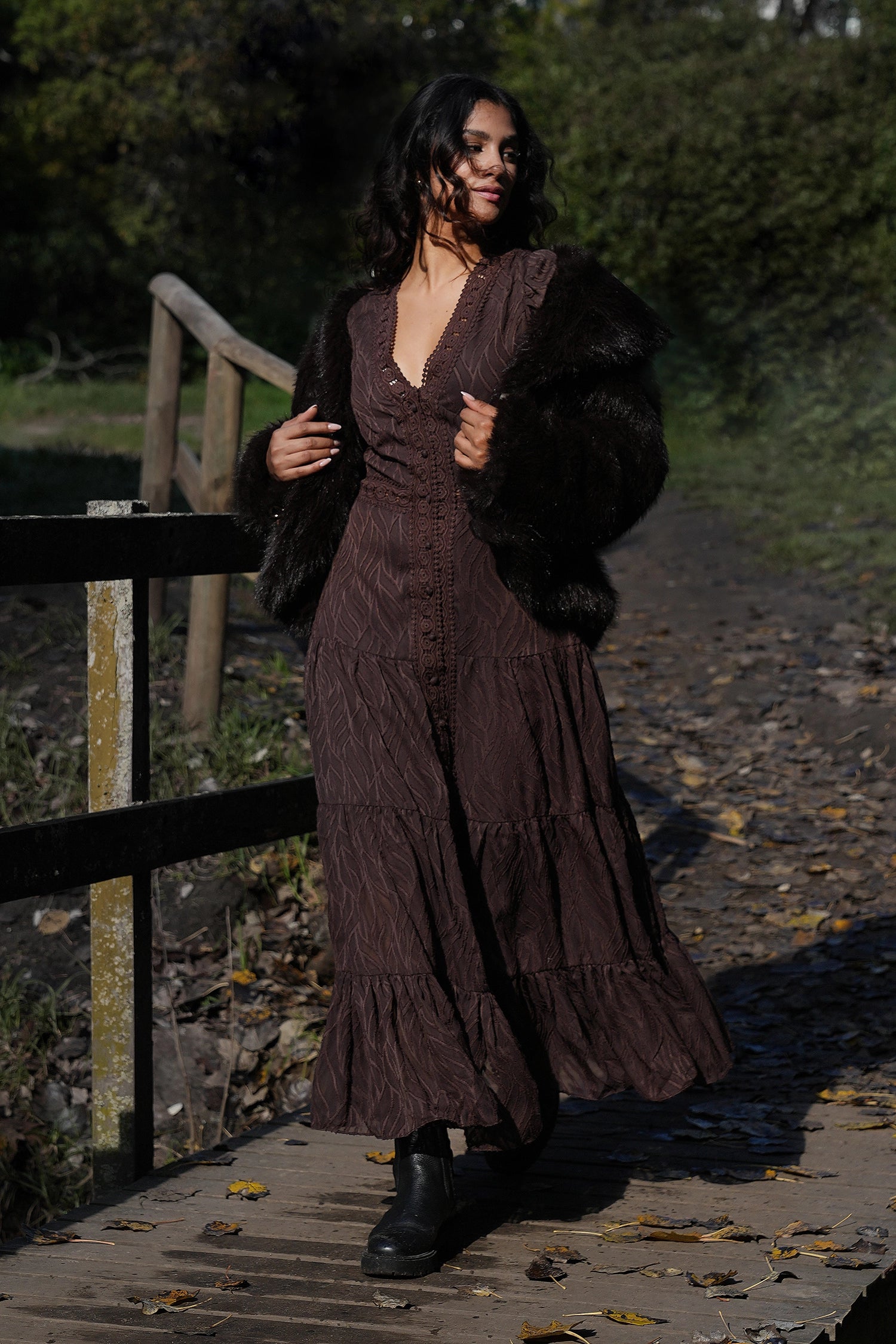 Woman in a long brown dress with a fur coat standing on a wooden path.