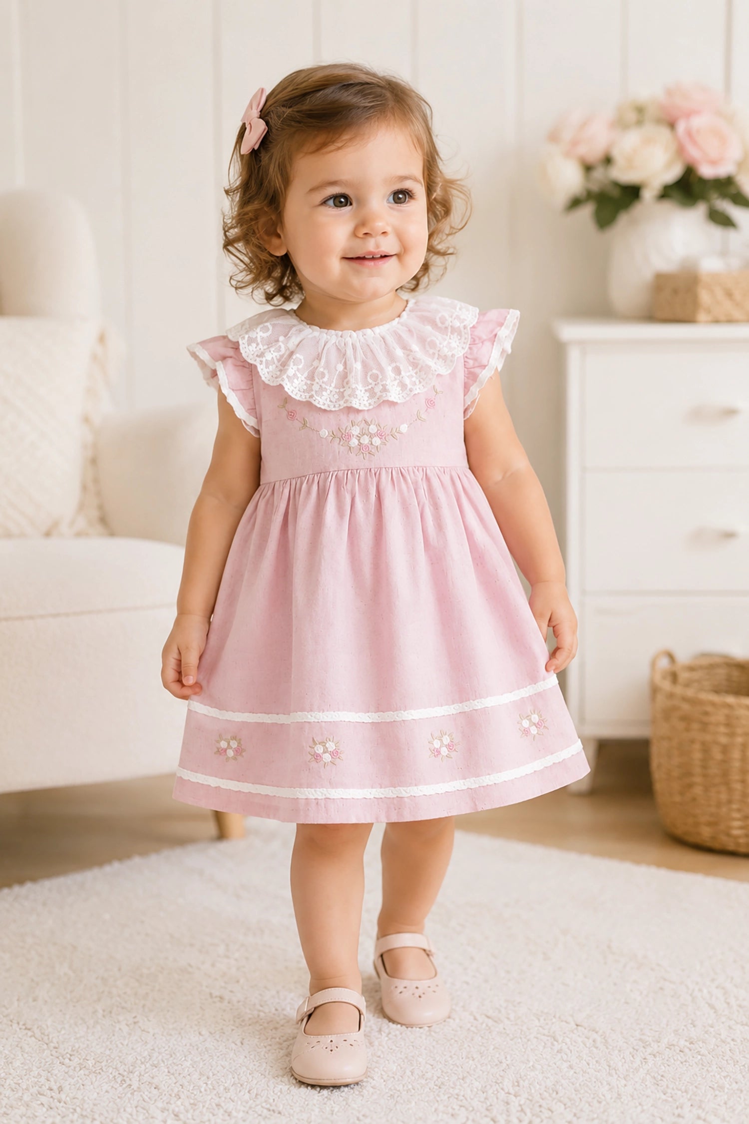 Young girl in a pink dress standing in a room with a couch and flowers in the background.