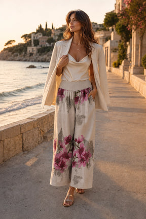 A woman stands on a seaside walkway at sunset wearing a cream-colored outfit with a floral print. The outfit consists of a sleeveless camisole top, a matching open-front blazer, and wide-leg pants featuring a pink and purple floral pattern over a cream base.
