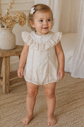 Baby in a lace romper standing in a softly lit room with a wooden stool and decorative flowers.