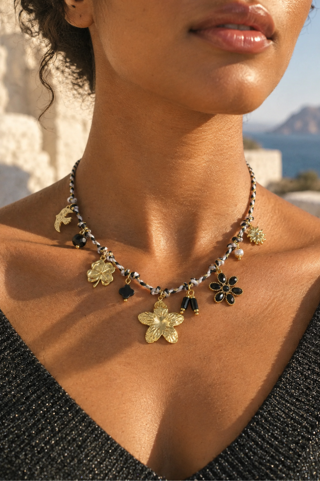 A close-up lifestyle shot of a model wearing a delicate gold and silver chain necklace adorned with floral charms, black beads, and white pearls, set against a sunny outdoor background.