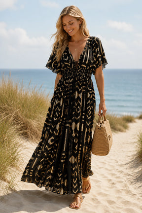 A woman wearing a black and beige geometric print maxi dress walks along a sandy beach path with the ocean and dunes in the background, holding a woven straw handbag.