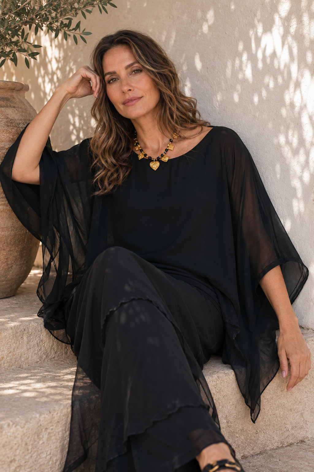 A woman wearing a black sheer long-sleeve tunic top and matching black pants, seated on stone steps against a textured wall with dappled sunlight.