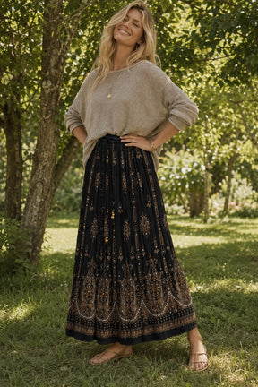 A woman standing outdoors in a grassy wooded area, wearing a beige knit long-sleeve top and a long black maxi skirt with an ornate gold-toned bohemian pattern.