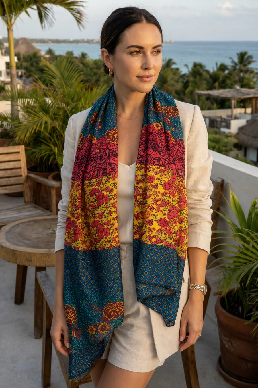 A woman wearing a cream-colored outfit and a long, multicolored patterned scarf with blue, red, and yellow floral sections, standing on a tropical balcony overlooking the ocean.