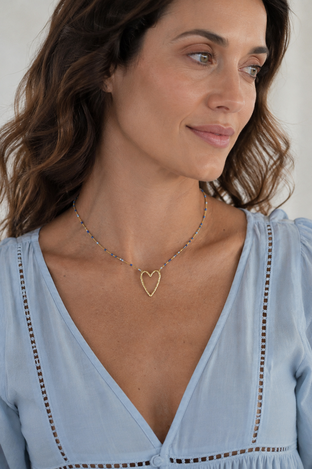 A close-up of a woman wearing a delicate gold heart pendant necklace over a light blue blouse.
