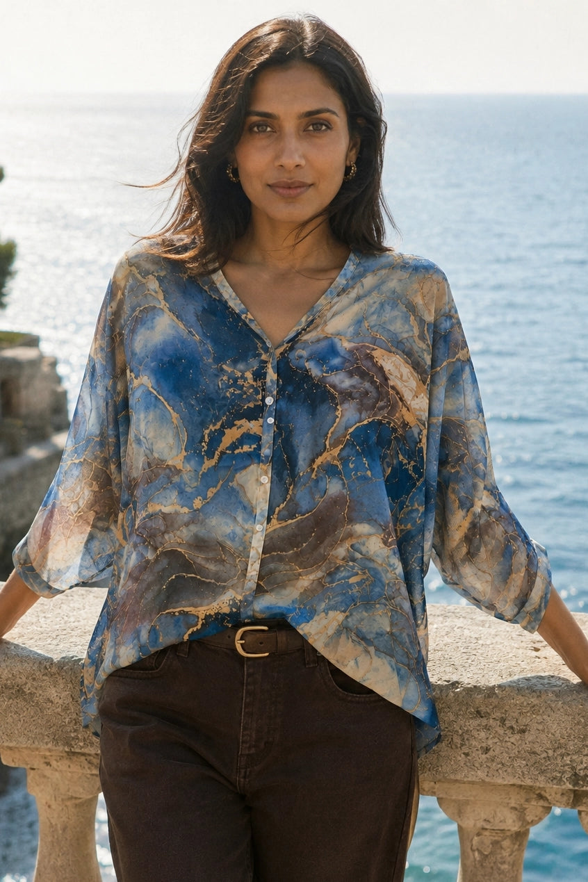A woman stands outdoors by the sea wearing a loose, semi-sheer blouse with an abstract blue, beige, and brown marbled print, paired with dark brown pants.