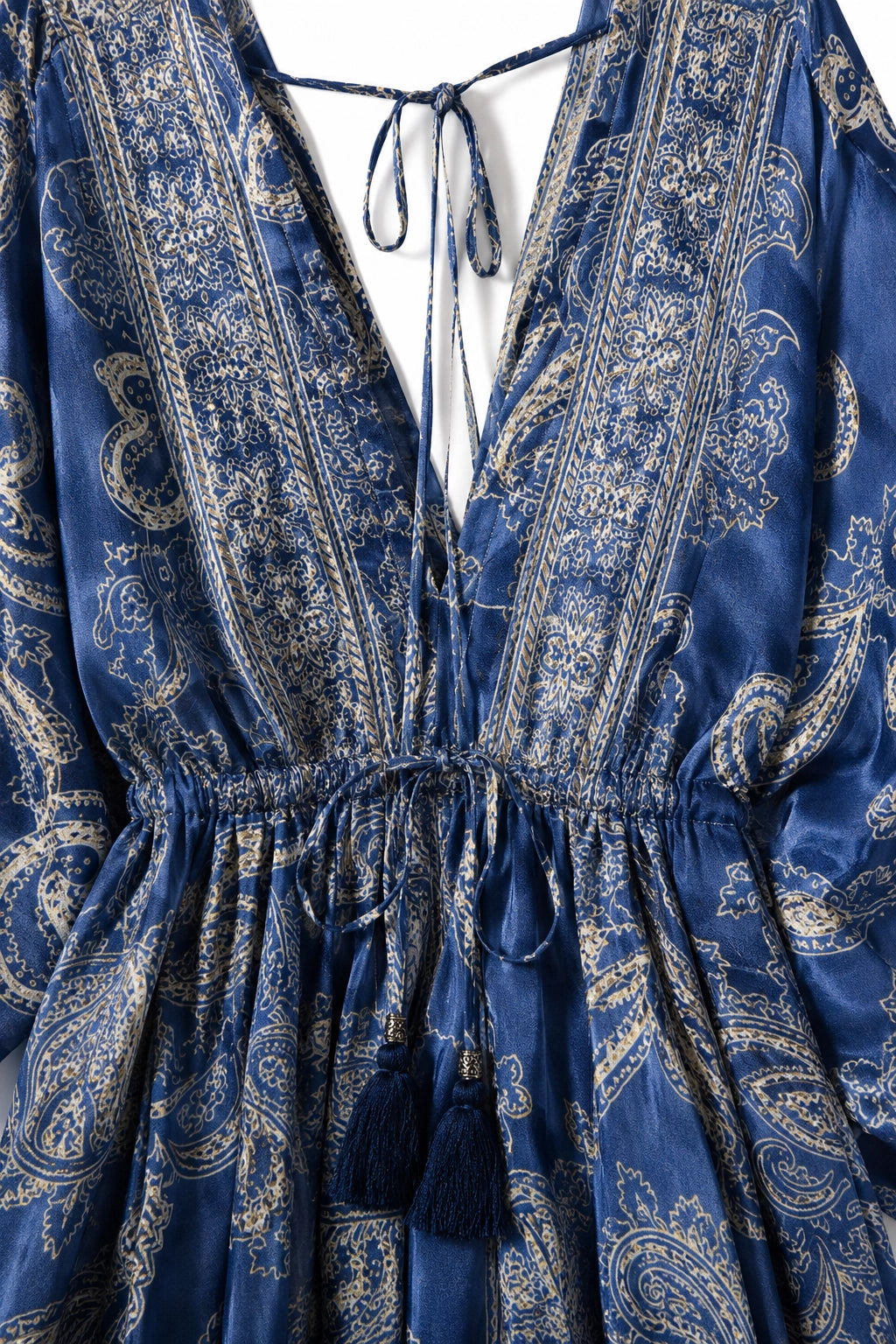 A close-up of a blue patterned dress with a deep V-neckline, tie details at the collar, a gathered elastic waist, and dark blue tassels hanging from the front.