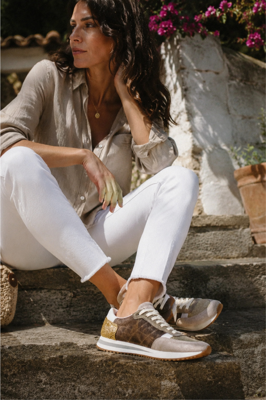 A woman sits on rustic stone steps outdoors, wearing a beige button-up shirt, white pants, and brown monogram sneakers with gold glitter accents.
