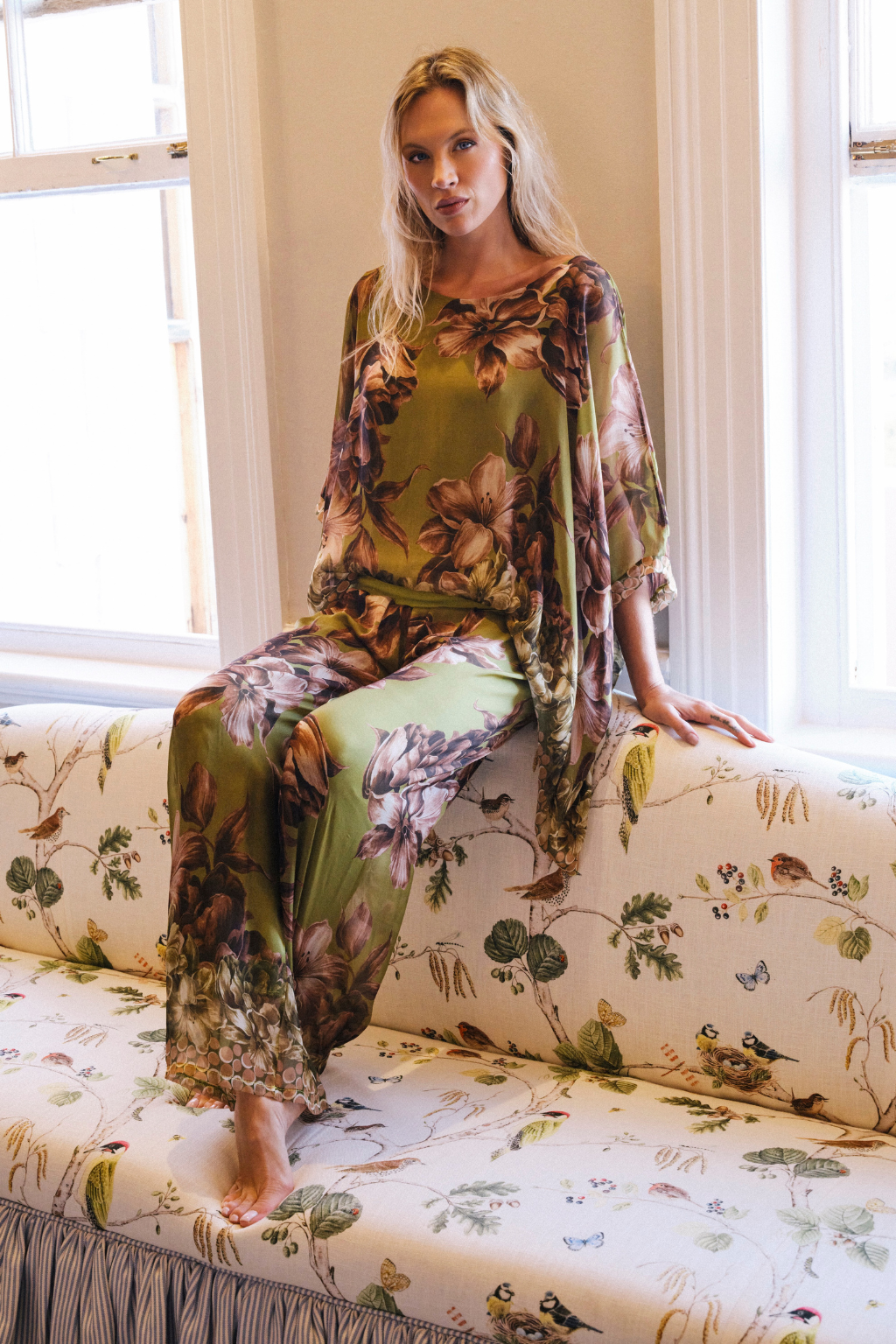 A woman sits on a floral upholstered bench in a bright room, wearing a matching olive green two-piece set with a large brown and beige floral print. The set includes a loose top and wide-leg pants, with a matching sheer drape.