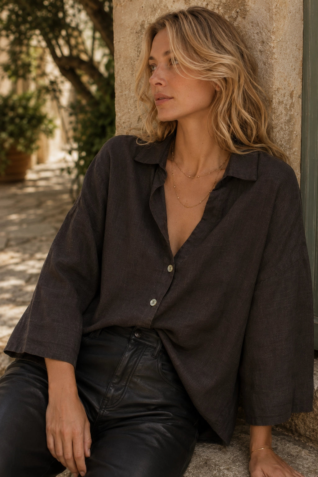 A woman wearing an oversized dark brown linen button-up shirt and black leather pants, leaning against a stone pillar in an outdoor setting.