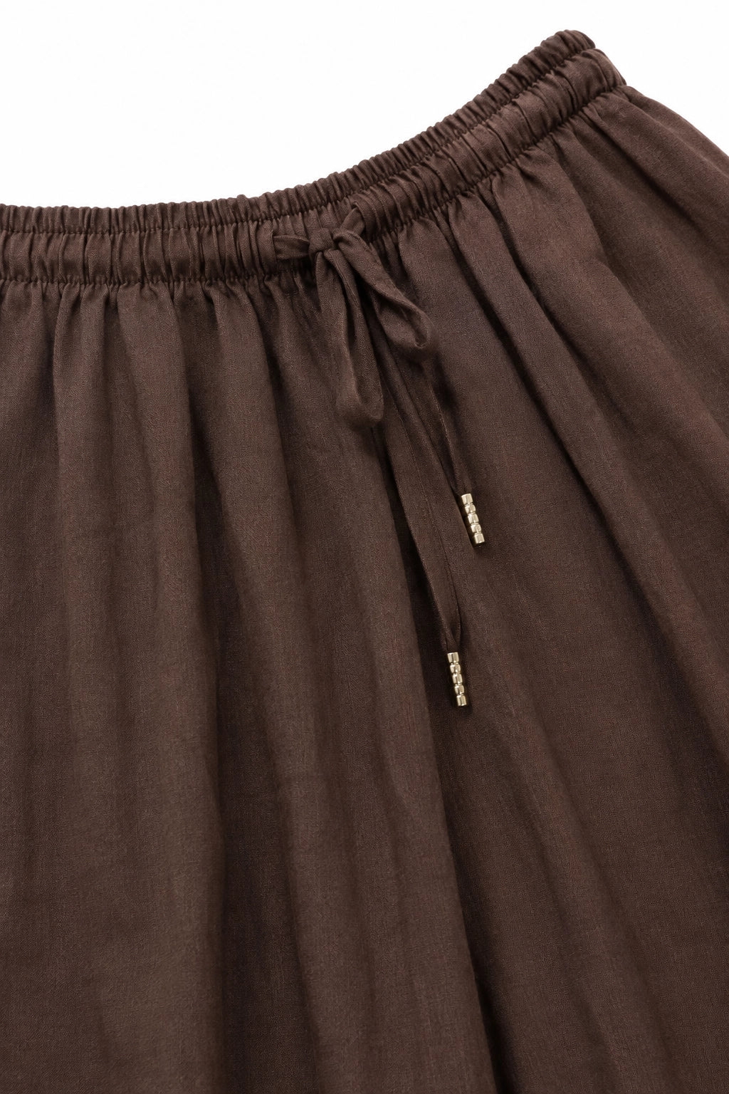 Close-up of a dark brown pleated skirt with an elasticized waistband and drawstring tie, featuring small gold-tone metal accents on the drawstring.