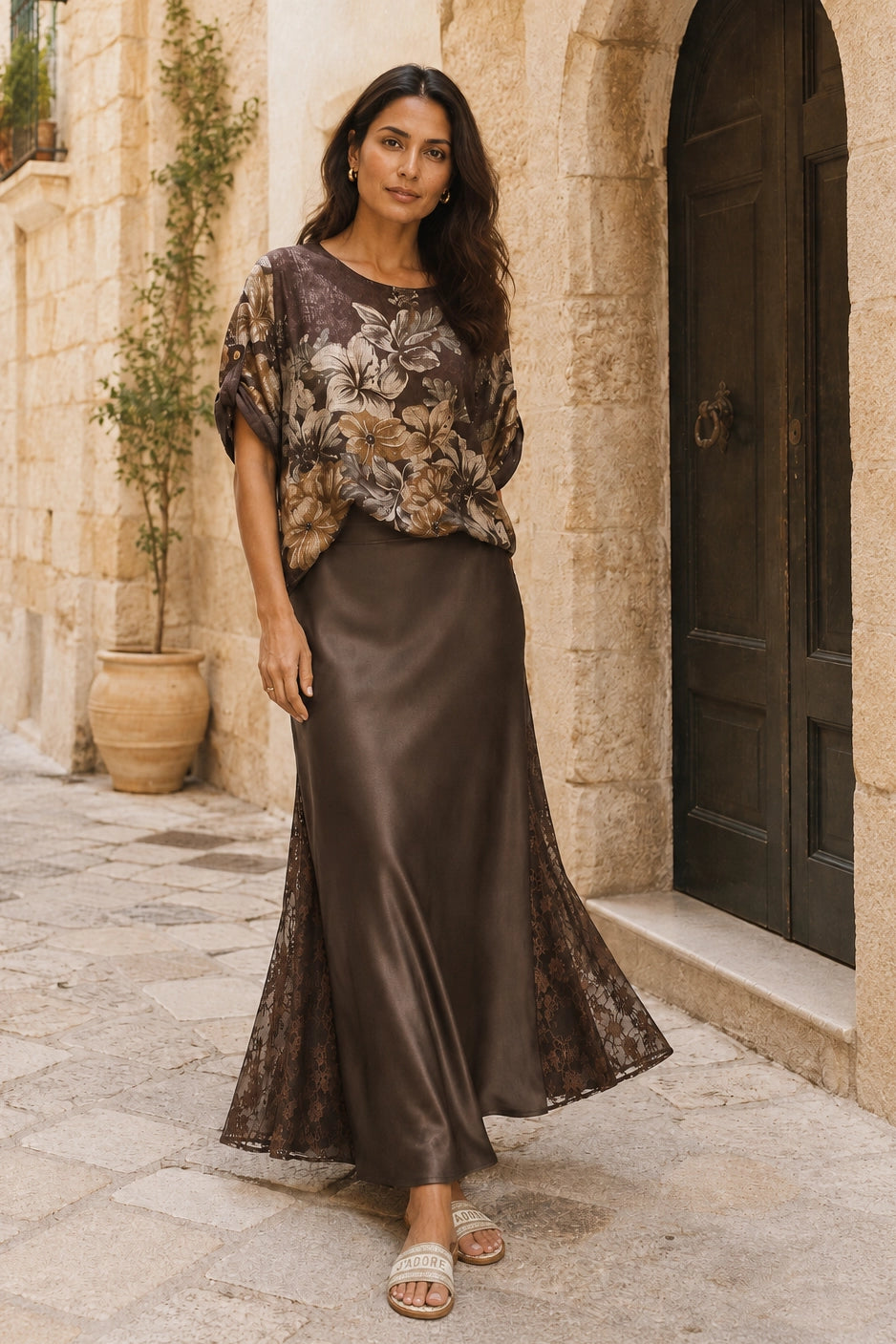 A woman stands on a stone walkway in front of a beige stone building with a dark wooden door, wearing a brown floral short-sleeve top and a matching brown satin maxi skirt with lace trim.