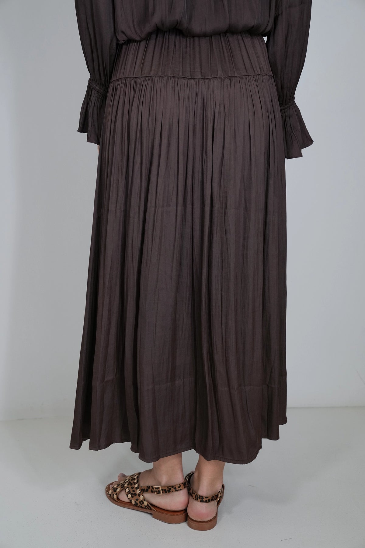 A dark brown pleated maxi skirt worn by a model, paired with leopard-print flat sandals, shown from the waist down against a plain light gray background.