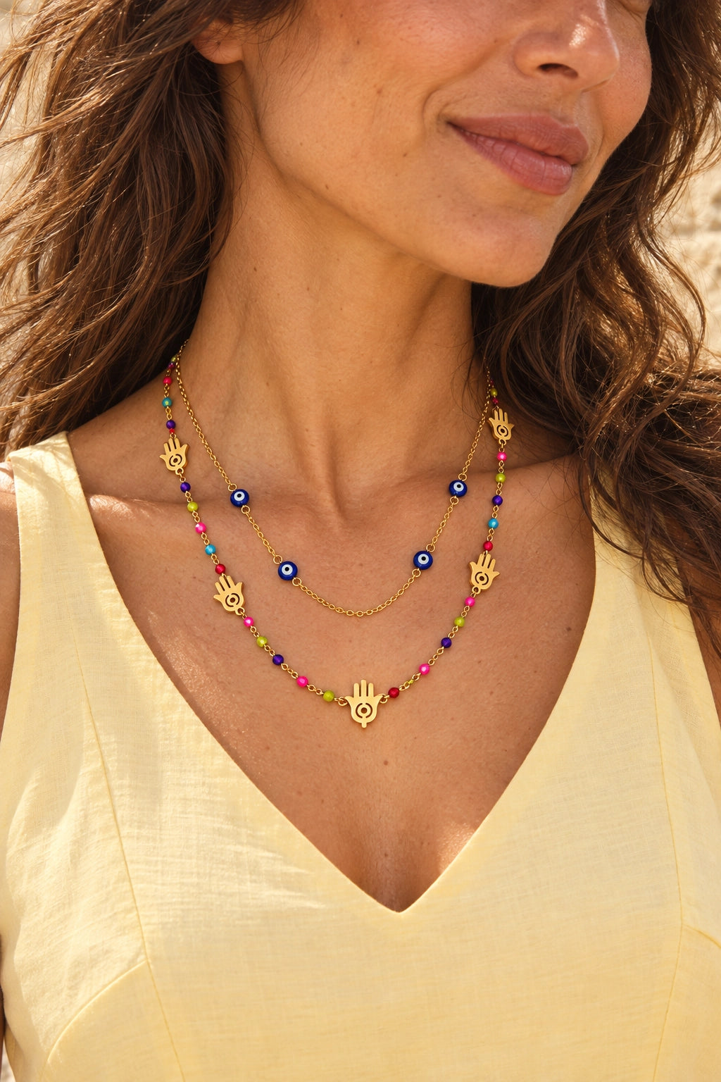 A close-up lifestyle shot of a woman wearing a layered gold necklace set. The top layer features gold Hamsa hand charms and evil eye beads, while the bottom layer is a gold Hamsa pendant with colorful seed beads in pink, blue, and green.