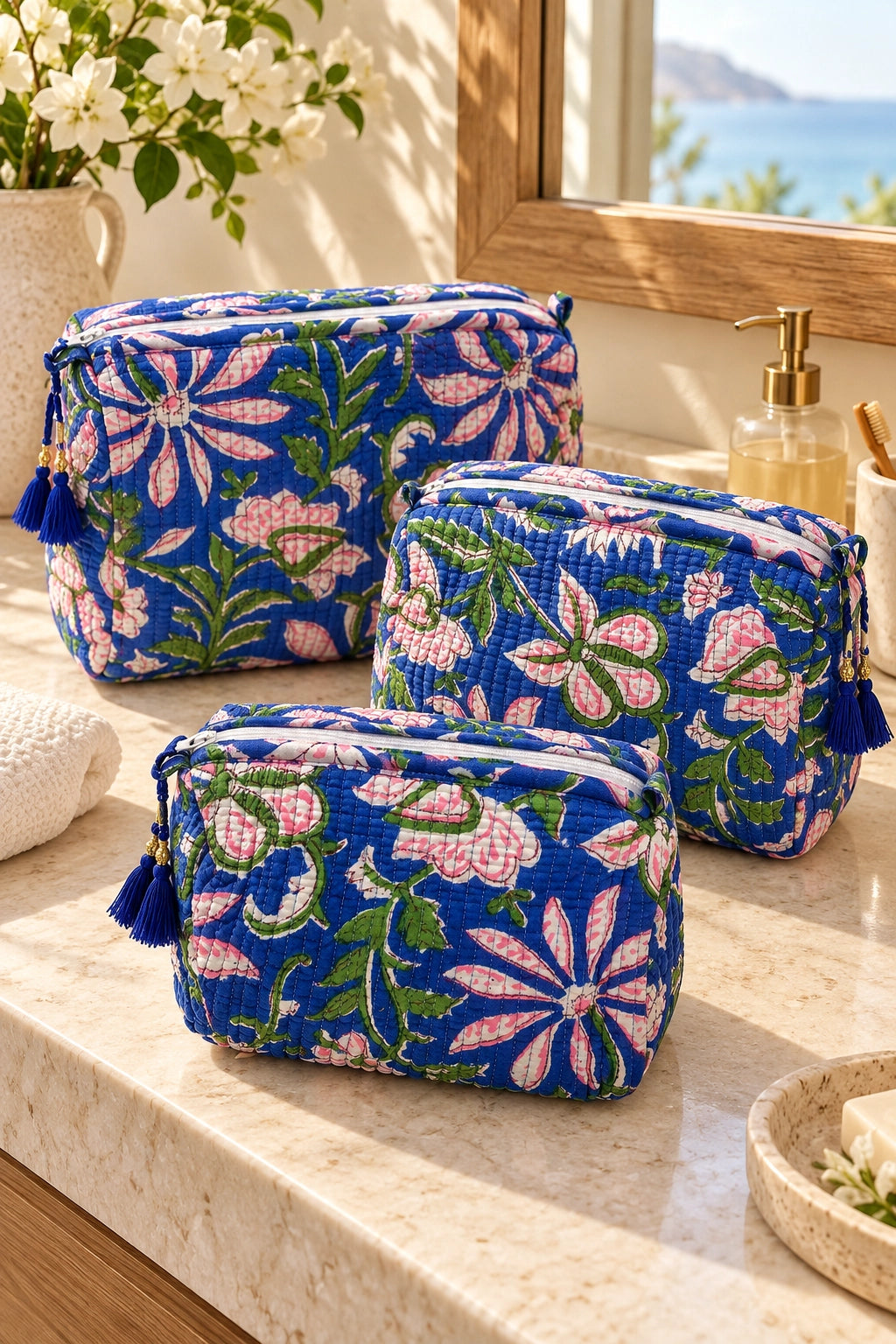 Three blue floral makeup pouches with tassel zippers arranged on a marble bathroom counter near a window.