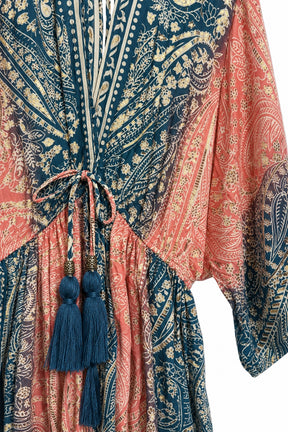 A close-up of a bohemian-style kaftan dress featuring a split design with navy blue and coral pink paisley patterns, cream vertical stripes, and a drawstring waist with blue tassels.