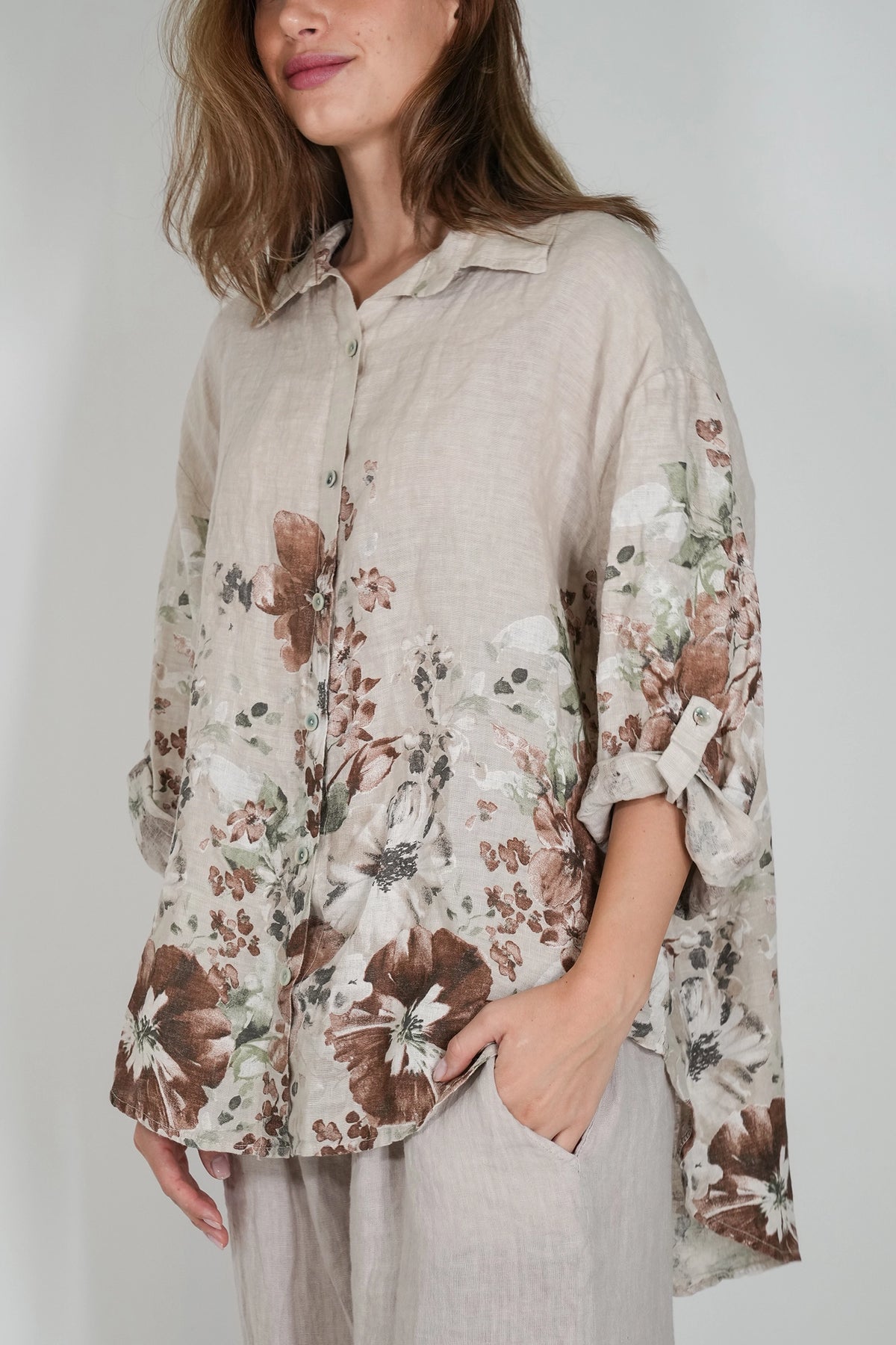 A woman wearing a loose, light beige linen button-up blouse with a brown and white floral print and rolled sleeves, paired with light gray trousers, against a plain light gray background.