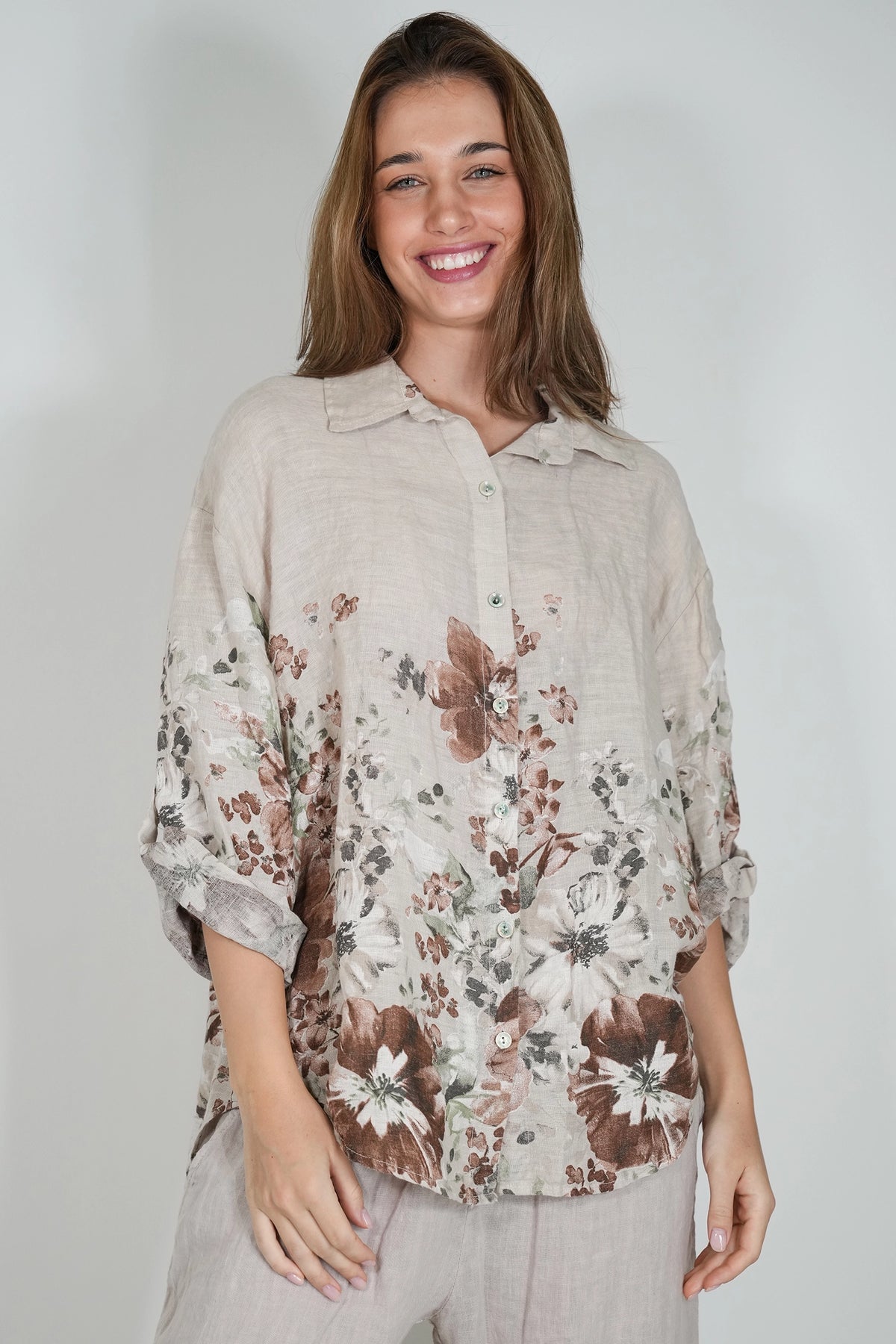 A woman wearing a light beige linen button-up shirt with a brown and white floral print, paired with light gray trousers, photographed against a neutral light gray background.
